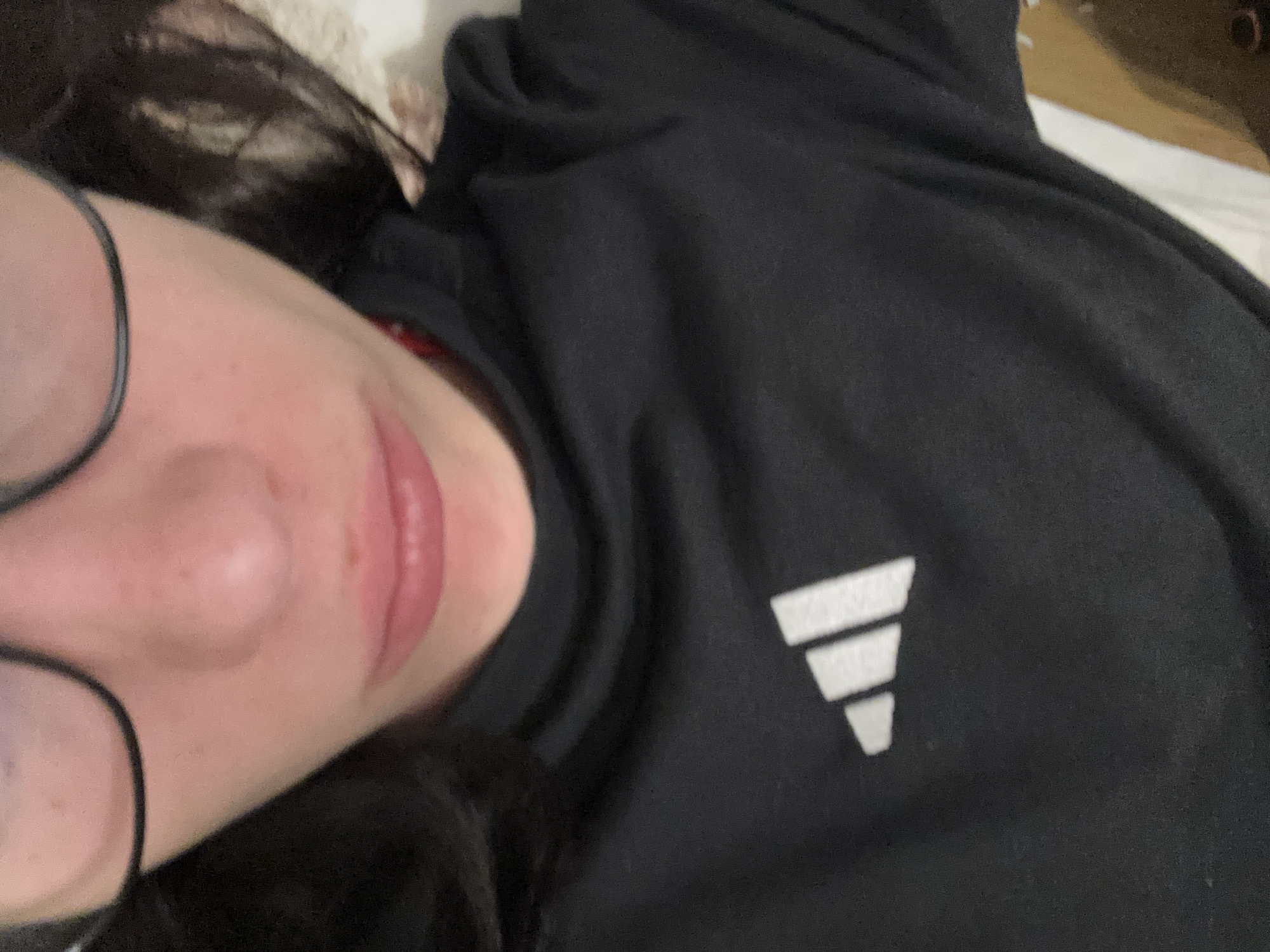 A close-up, partially obscured selfie of a person with fair skin, wearing glasses, and a black Adidas sweatshirt. The person's nose, lips, and part of their left eye (through the glasses) are visible. Part of the person's dark hair can be seen. The Adidas logo is visible on the sweatshirt. The background includes part of a white or light-colored surface, potentially a bed or couch. Location information (Buenos Aires, Argentina) is not discernible from the image.