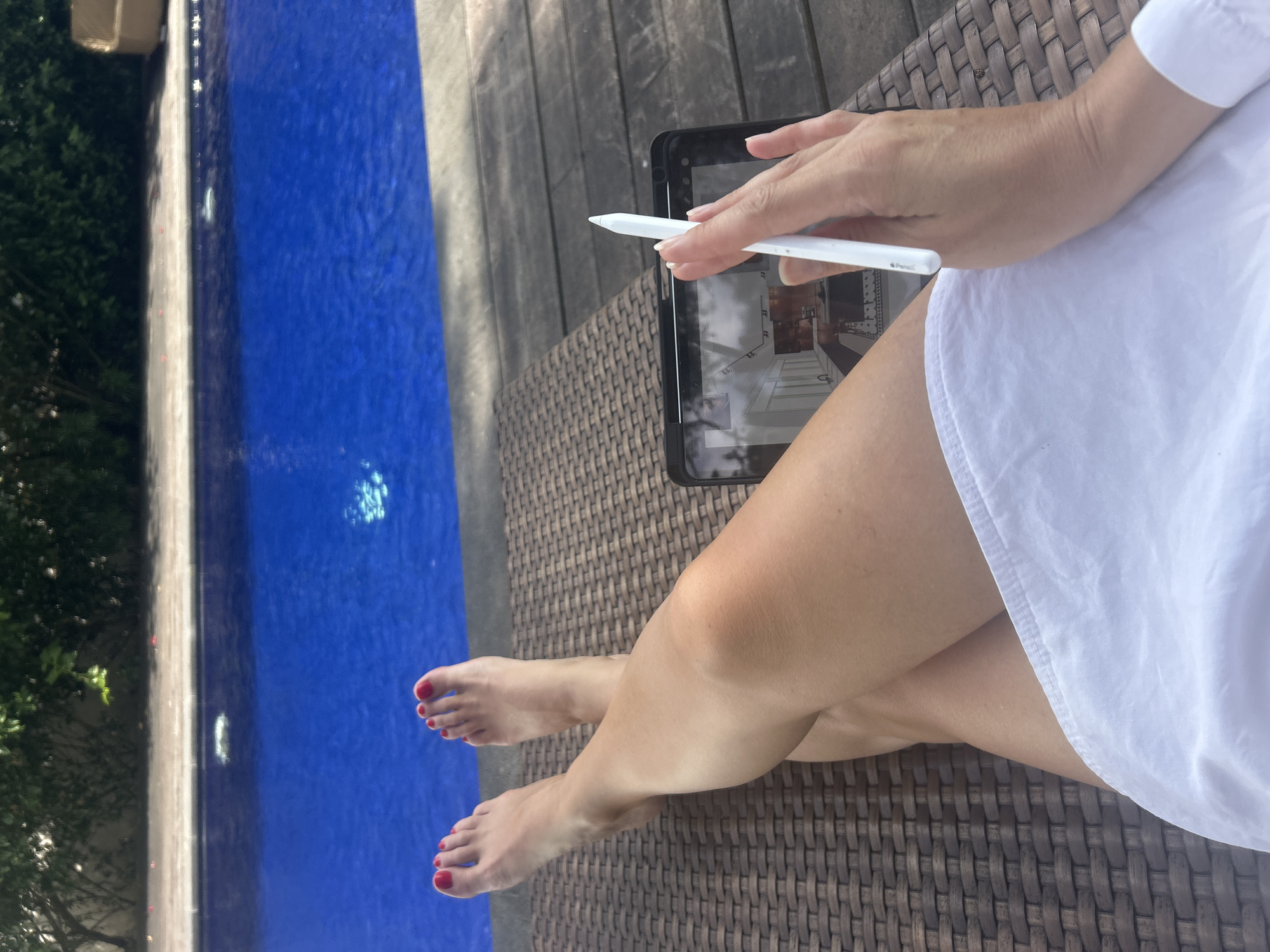 A woman sits in an outdoor chair near a pool, holding a white stylus above a tablet displaying a room design. Her legs are crossed, and her toes are painted red. The setting includes a wooden deck and glimpses of greenery. The location cannot be determined as Kediri, Indonesia, based on the provided image.