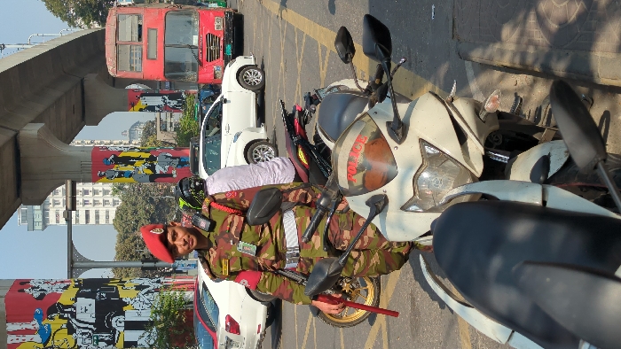 A man in a military uniform and red beret stands next to a parked motorcycle with "POLICE" written on its windshield. Cars and a red double-decker bus are visible in the background, under an elevated structure decorated with colorful street art. The scene suggests an urban environment with traffic and public transportation. The man is holding a red and gold stick, possibly related to traffic control.