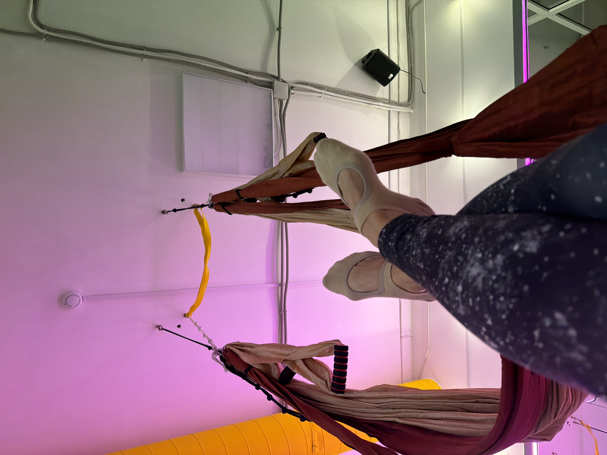 The image shows a person's legs and feet in soft ballet slippers, suspended in aerial silks. The silks are red and tan, with black handles. The background is a ceiling with pink and green lighting, electrical conduits, and a smoke detector. There is also a yellow tube and a black speaker mounted on the ceiling. The person is wearing dark leggings with white speckles. The overall impression is of an aerial fitness or dance studio. There is no discernible information in the image to indicate the location in the city of Chaikovsky, Russia.