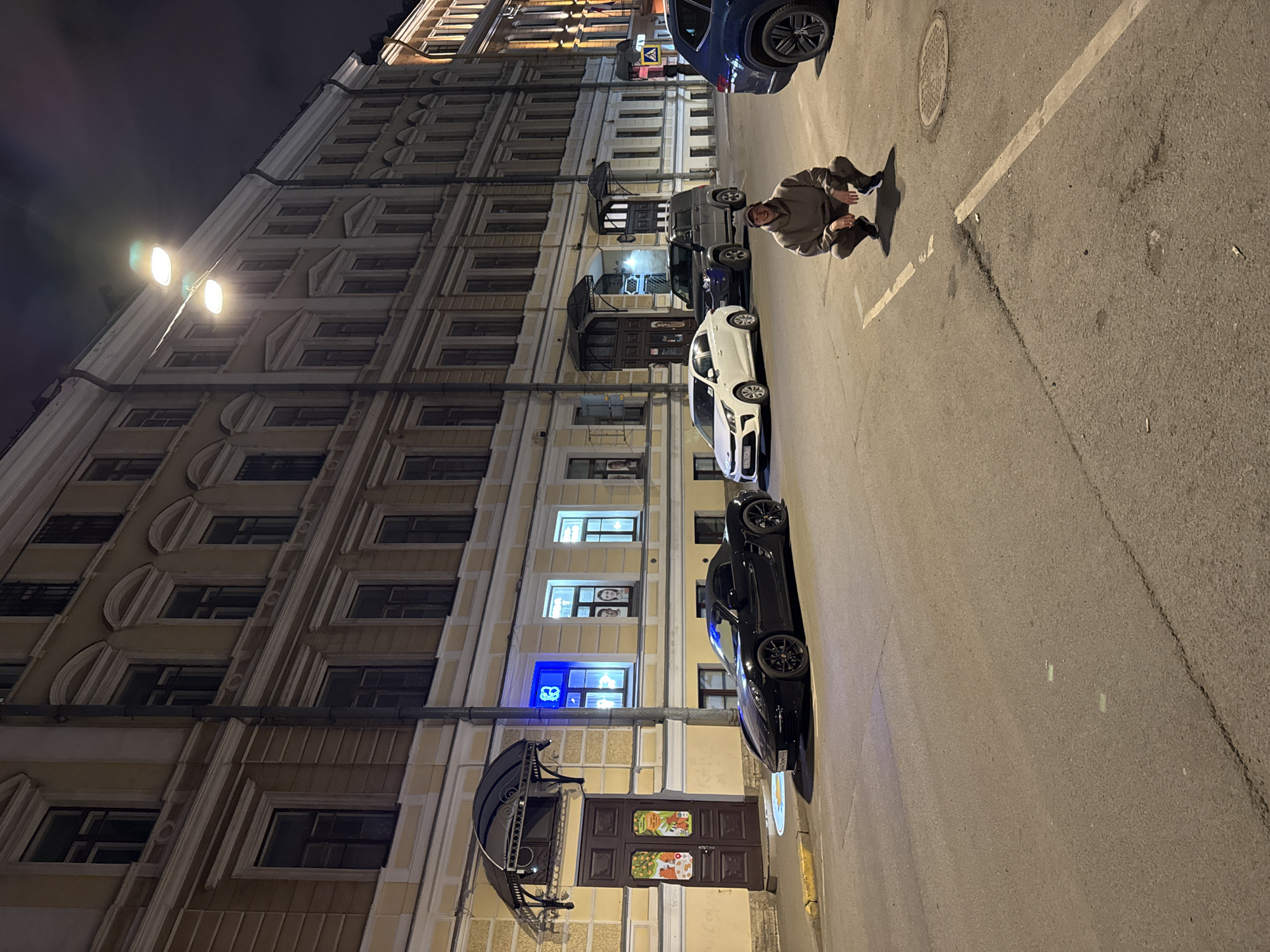 The image depicts a street scene in Saint Petersburg, Russia at night. The street is illuminated by street lamps and artificial lighting from buildings. Buildings with classical architecture line the street. Several parked cars are visible, including a dark blue SUV, a silver SUV, a white sedan, and a black sports car. A person is crouched on the sidewalk near the parked cars, looking in the direction of the camera. The street surface appears to be asphalt. The overall scene suggests an urban environment at night.