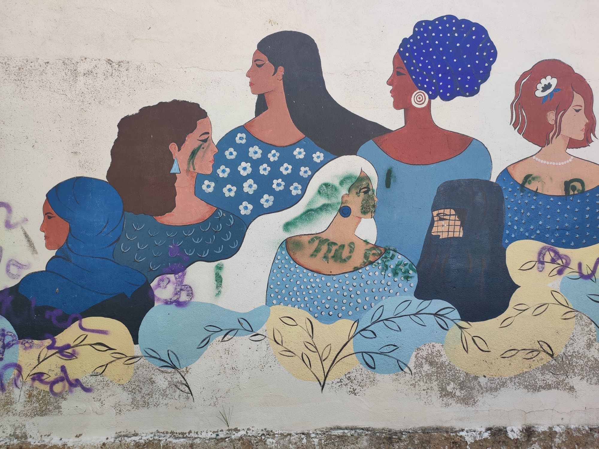 This is a mural painted on an outdoor wall in Traspinedo, Spain, depicting a group of diverse women. The mural features a row of women with varying skin tones, hairstyles, and head coverings, including a woman in a blue hijab and another in a blue patterned turban. Several women have green markings on their faces, resembling war paint or makeup. Their clothing is primarily in shades of blue, with some featuring floral or polka dot patterns. The women are depicted from the shoulders up, looking in different directions. The mural is set against a distressed white wall with visible cracks and discoloration. Below the figures, there are abstract shapes in yellow and light blue, with black line drawings of leaves and branches. Some of the lower sections of the mural show signs of graffiti, with purple spray paint marks.