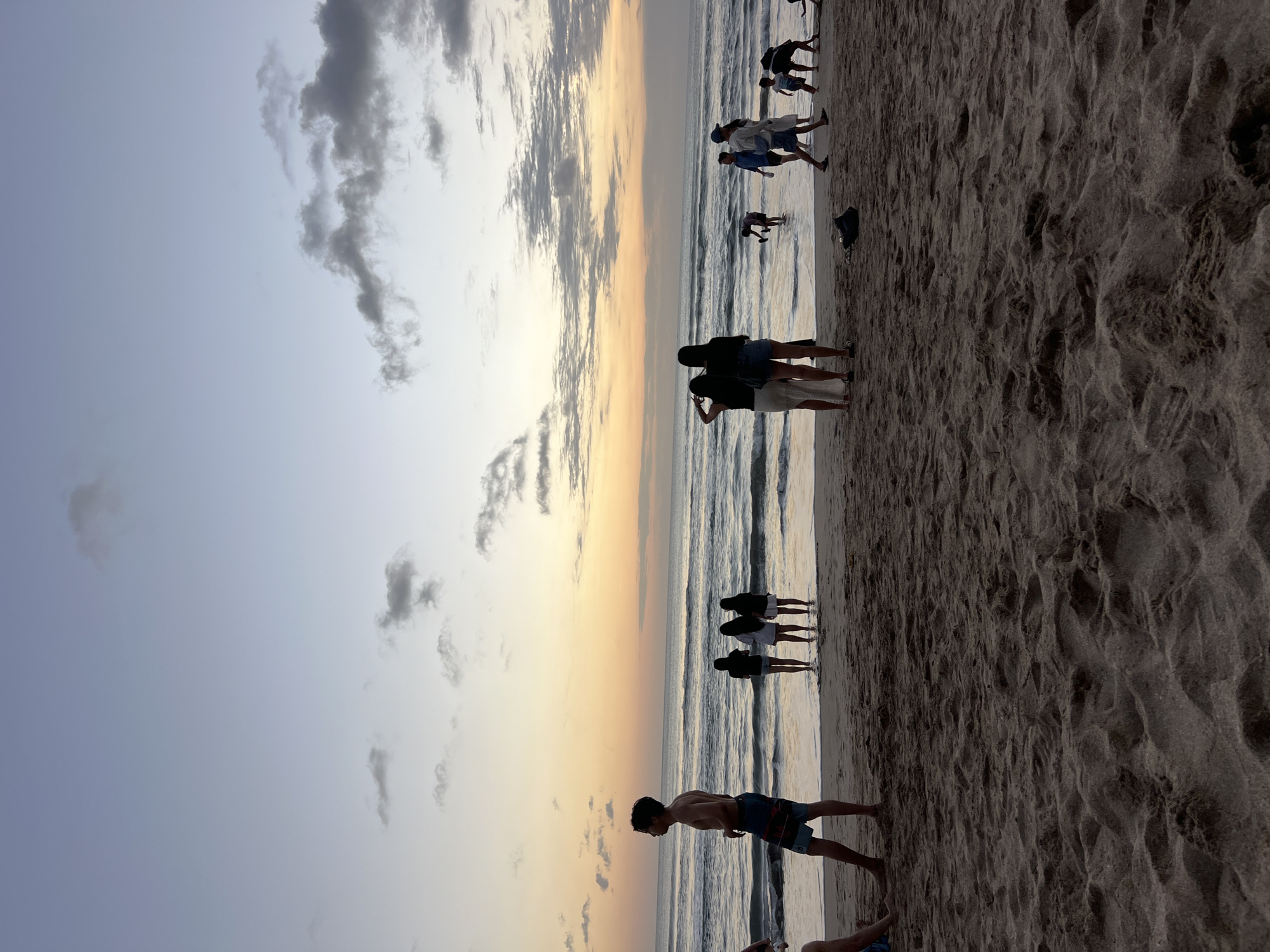 The image shows a beach in Kuta, Indonesia, during sunset. The sky is a gradient of soft blues and oranges, with scattered clouds. The ocean is calm with gentle waves rolling onto the sandy shore. Several people are present on the beach, some wading in the shallow water, others walking or standing on the sand. In the foreground, the sand is textured with footprints and ripples. In the mid-ground, a group of people stand near the water's edge, silhouetted against the sunset. Further back, more individuals are visible near the ocean. The overall atmosphere is serene and casual, capturing a typical evening scene at a popular beach destination.