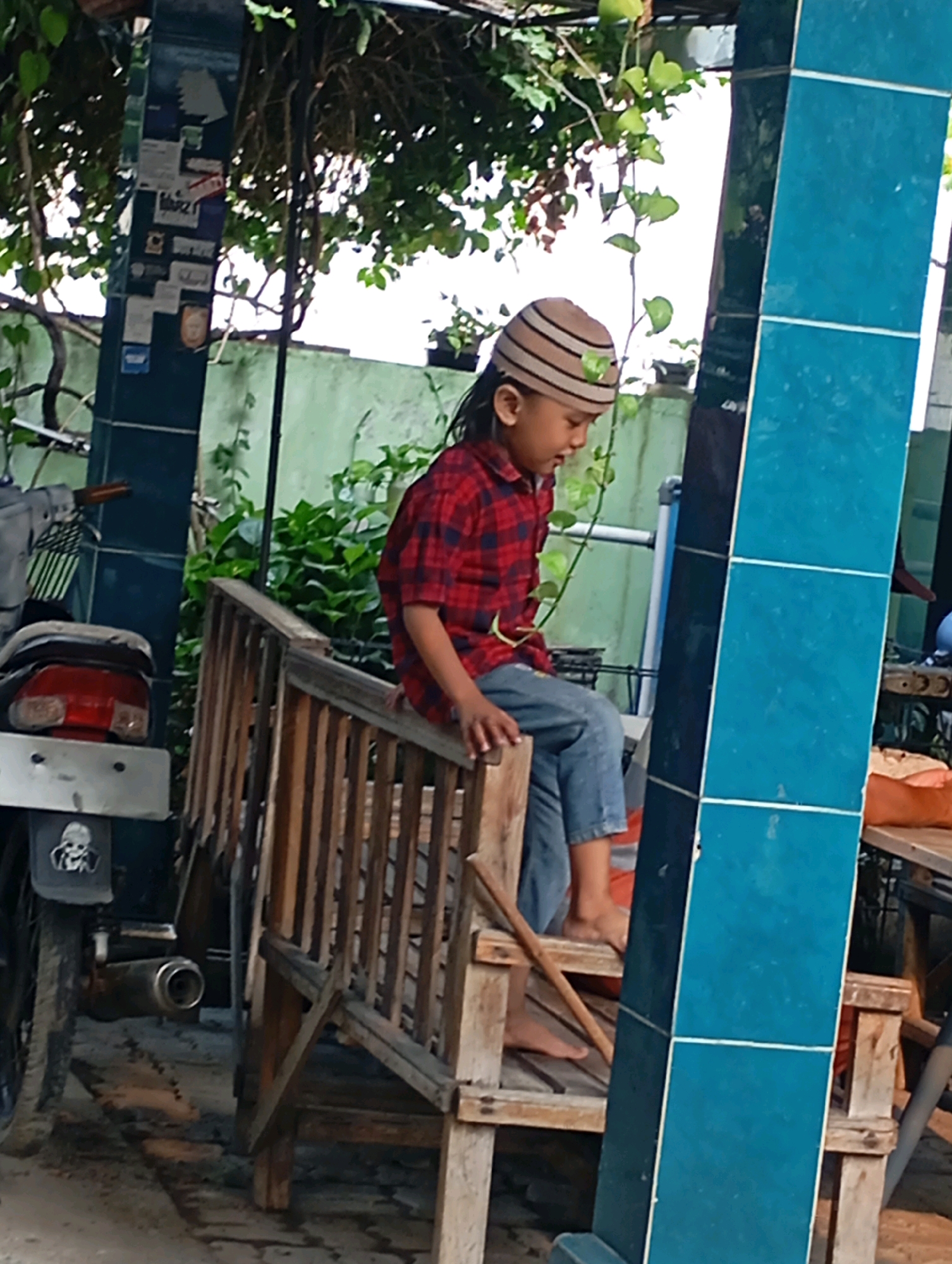 The image depicts a young child, likely male, wearing a red and black plaid long-sleeved shirt, blue jeans, and a striped knit cap, sitting on a wooden bench. The child is looking down, with their head tilted to the side. To the left of the child, a motorcycle is partially visible, with its rear taillight and exhaust pipe discernible. The bench appears to be made of rough-hewn wood and is positioned outdoors, with greenery and trees in the background. A large, blue tiled structure is prominent on the right side of the image. The setting appears to be a casual, outdoor area, possibly a yard or a communal space in Babelan, Indonesia, given the context provided.