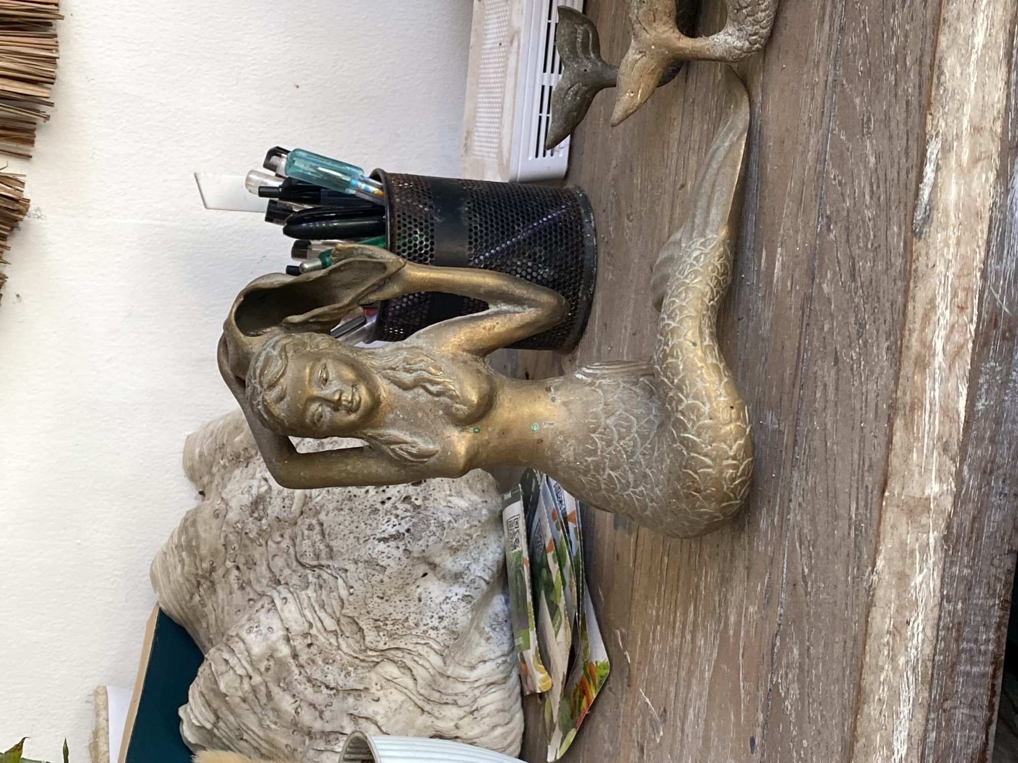 An antique brass mermaid figurine with a weathered patina sits on a distressed wooden table. The figurine depicts a mermaid with her tail curled to the right and her arms raised, holding a large shell over her head. Behind the mermaid, a textured, large shell object is visible, possibly a giant clam shell. To the right of the mermaid, a black mesh cup is filled with pens and markers. Further to the right, a white electronic device with a perforated surface is placed on the table, and a small brass fish tail sculpture is positioned in front of it. The background consists of a white wall, with dried palm fronds hanging from the ceiling. The overall scene suggests a coastal-themed indoor display. The location is Kuta Selatan, Indonesia.