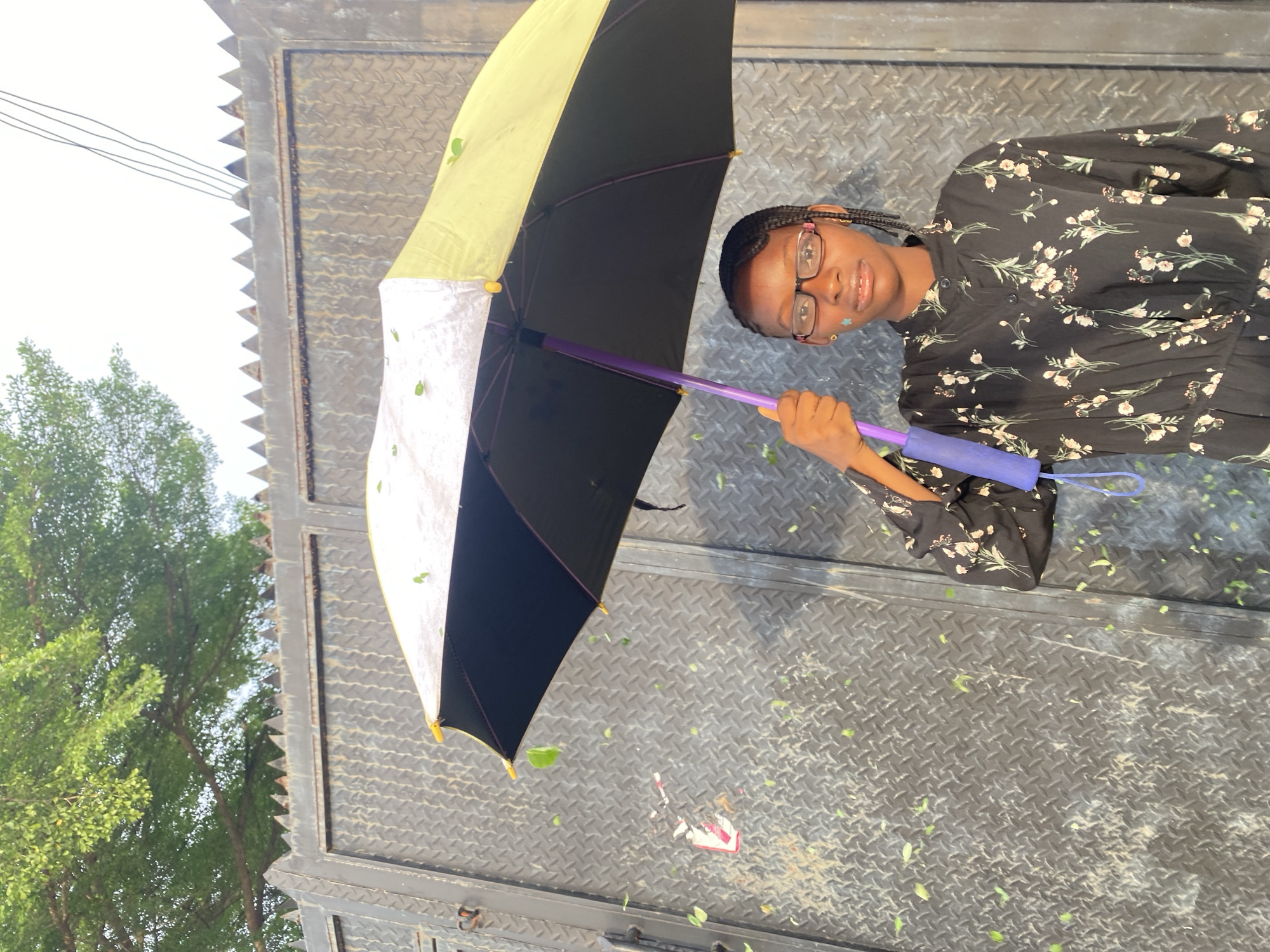 A young female stands outdoors in the city of Iranla, Nigeria, positioned in front of a dark, heavy-duty metal gate featuring a diamond plate pattern and topped with triangular anti-climb spikes. The female, appearing adolescent, has dark skin, braided hair, and wears eyeglasses with pink frames. A small light-colored mark is visible on her left cheek. She is dressed in a dark-colored, long-sleeved garment with a white or light-colored floral pattern, cinched at the waist. She holds an open umbrella in her right hand, gripping its purple shaft; the umbrella canopy is two-toned, featuring a lighter upper section and a darker lower section, with a blue strap attached to its handle. Numerous small green leaves are airborne and resting on the umbrella's surface and the metal gate. A tree with green foliage is visible in the upper left background, while thin utility wires traverse the upper right against a bright, overcast sky. A small, faded, and partially torn sticker is affixed to the metal gate to the left of the subject. The female looks directly towards the camera with a neutral expression.