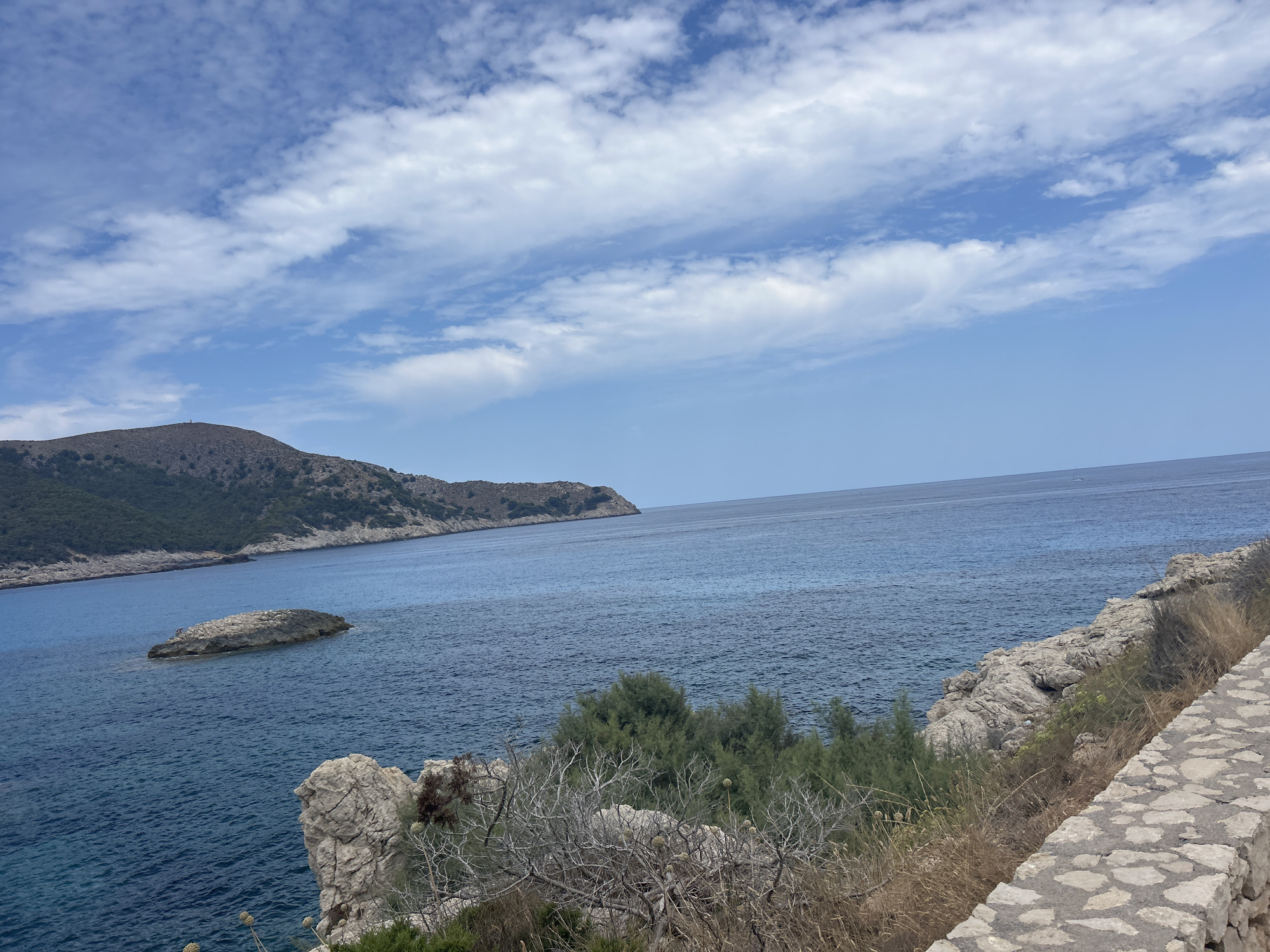 The image depicts a coastal landscape with the sea and a hilly coastline under a partly cloudy sky. In the foreground, a paved pathway runs along the right edge, constructed with irregular stones. Adjacent to the path, the terrain is rocky with dry vegetation and some sparse green shrubs.
The sea, a deep blue, dominates the middle ground and extends to the horizon. A small, rocky islet is visible in the sea to the left. Further in the distance, a mountainous coastline forms the backdrop, covered in vegetation and rising to a peak. The sky above is blue with scattered white clouds.
This scene is located in Cala Lliteres, Spain. There are no people or animals visible in the image, and no specific actions are being depicted. The focus is on the natural environment and the coastal infrastructure.