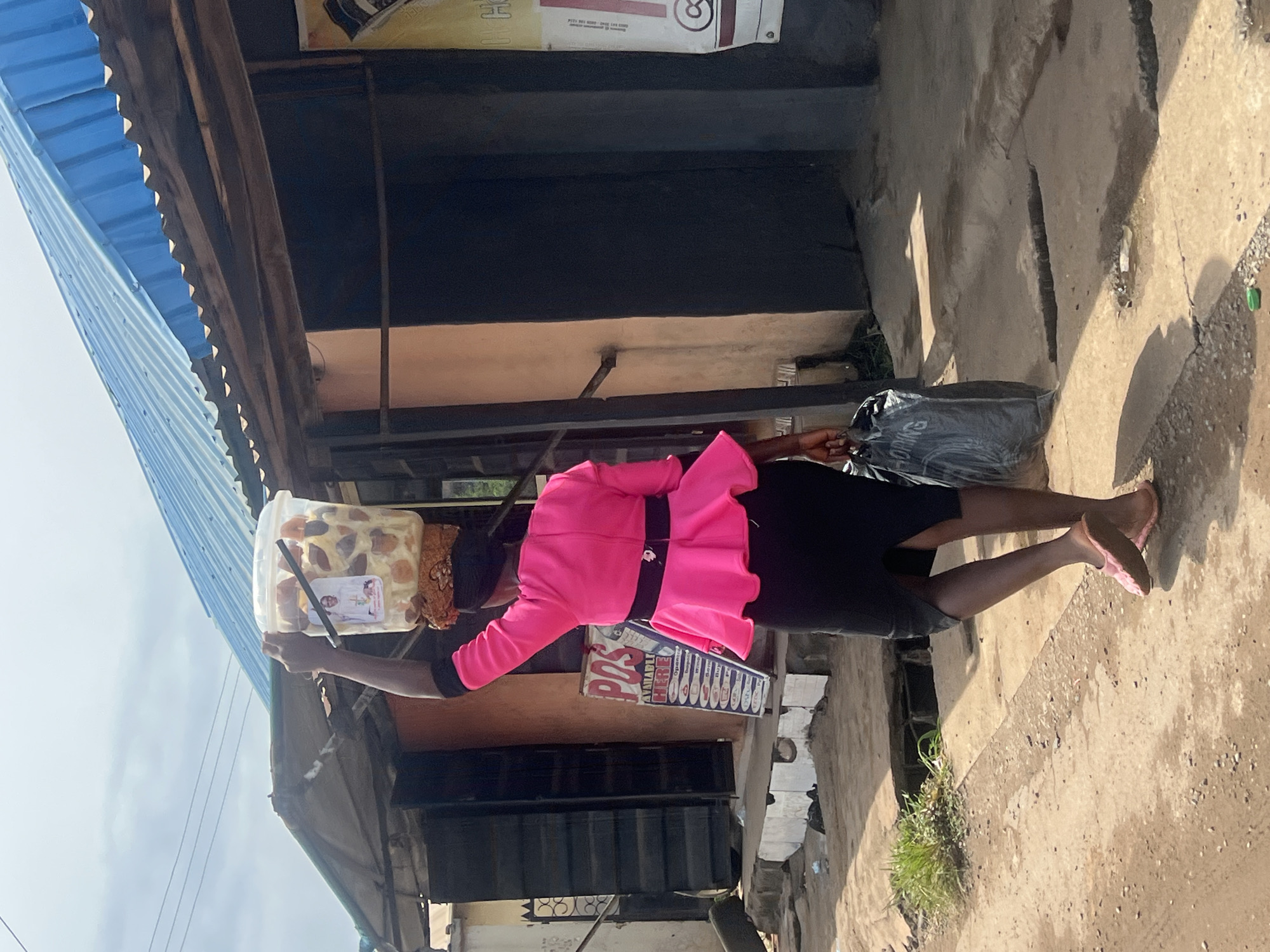 A woman walks along a dirt path in Nkpor, Nigeria, carrying a bucket of goods on her head and a black plastic bag in her hand. She is wearing a bright pink peplum top with black trim and a black skirt. Her feet are bare, clad in pink flip-flops. The path is dusty and cracked, with patches of green grass and debris visible. In the background, a corrugated blue roofed structure with wooden supports and a weathered facade suggests a commercial area or marketplace. A sign advertising "POS AVAILABLE HERE" is partially visible on the wall of one of the buildings. The overall scene depicts a common street vendor scenario in an urban environment.