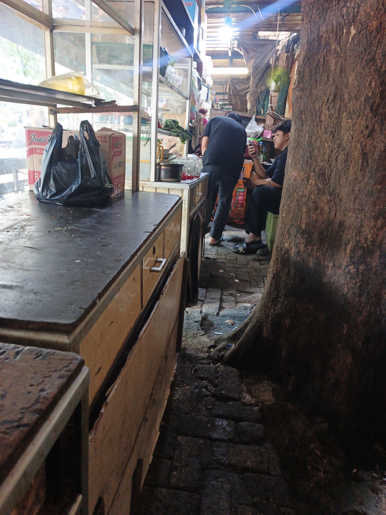 Two young men are seen at a roadside stall in South Jakarta, Indonesia. One man, wearing a black t-shirt and dark pants, is seated on a green cushion, looking at his phone and holding a beverage. The other man, in a black t-shirt and dark pants, stands facing away from the camera, also holding a beverage. The stall features shelves with various items, including food packaging like "Indomie" noodles, and two black plastic bags. A large tree trunk is prominent in the foreground. The ground is paved with bricks, and there are some scattered items on the ground. The scene appears to be an outdoor food or convenience stall.