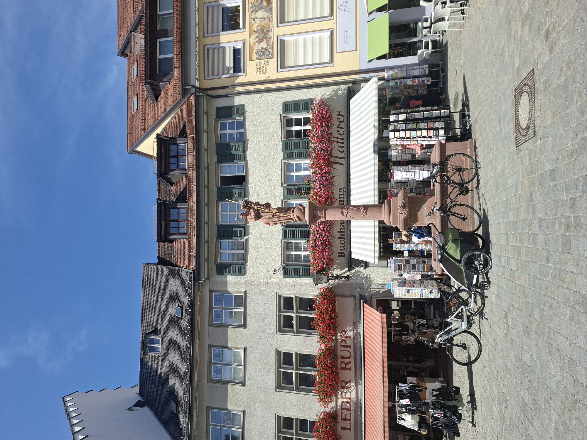 The image depicts a sunlit cobblestone plaza in Wangen im Allgäu, Germany. In the center, a prominent stone fountain features a statue of a robed figure holding a child, adorned with cascading red and pink flowers. To the left of the fountain, a building displays signs reading "LEDBER RUPP" and "Buchhaus Natterer," suggesting retail establishments. An orange and white striped awning extends over the "LEDBER RUPP" shop.
In front of the shops, two bicycles are parked. One is a white electric bicycle with a green child trailer attached. The second bicycle is a black road bike. A rack of black and white handbags is displayed outside the "LEDBER RUPP" store. Next to the fountain, racks of postcards are arranged.
Several individuals are visible. One person, wearing a blue shirt and shorts, stands near the bicycle with the trailer. Another person, wearing a pink shirt, browses the postcards. A person in dark clothing is also visible near the postcard display. In the background, several white chairs and tables suggest an outdoor seating area for a cafe or restaurant. The architecture of the buildings features multi-story facades with windows and pitched roofs, characteristic of European city centers. The sky is a clear, bright blue with a few wispy clouds.