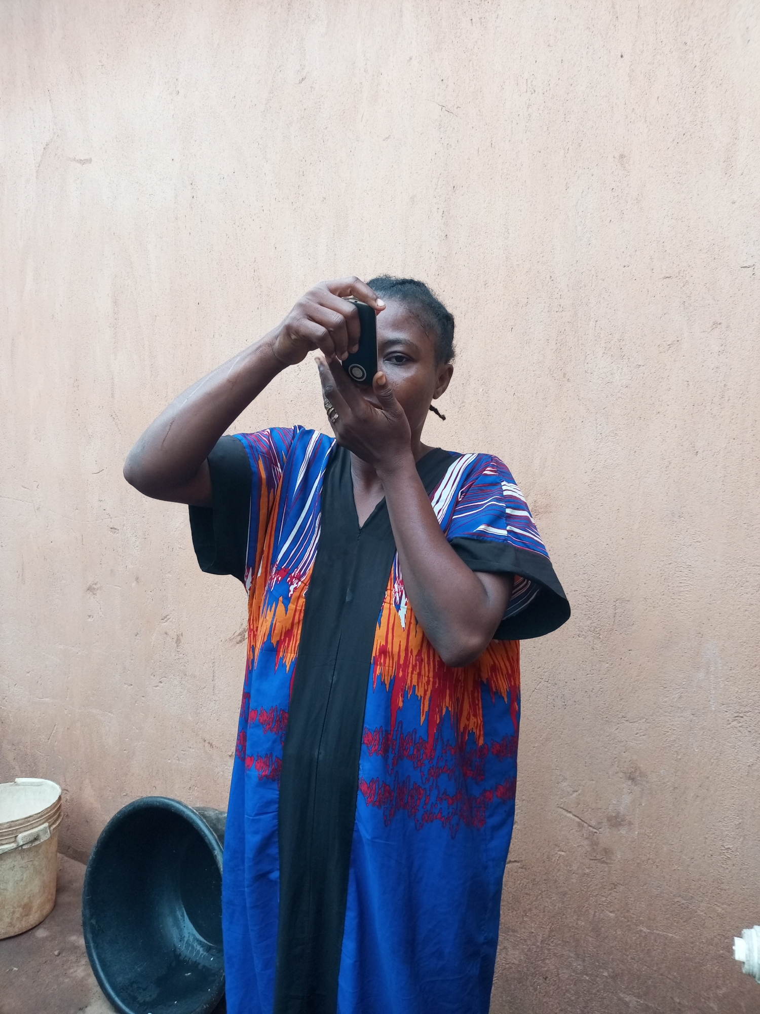 A woman stands in front of a textured, light brown wall, holding a small, black electronic device up to her eye as if taking a picture. She wears a loose, vibrantly colored robe with a black V-neck and a pattern of red, orange, and blue abstract designs. Her dark hair is pulled back. To her left, a beige plastic bucket and a large, black plastic basin are visible on the ground. The scene appears to be outdoors, possibly in a residential area. The location is not specified as Tempure, Nigeria.