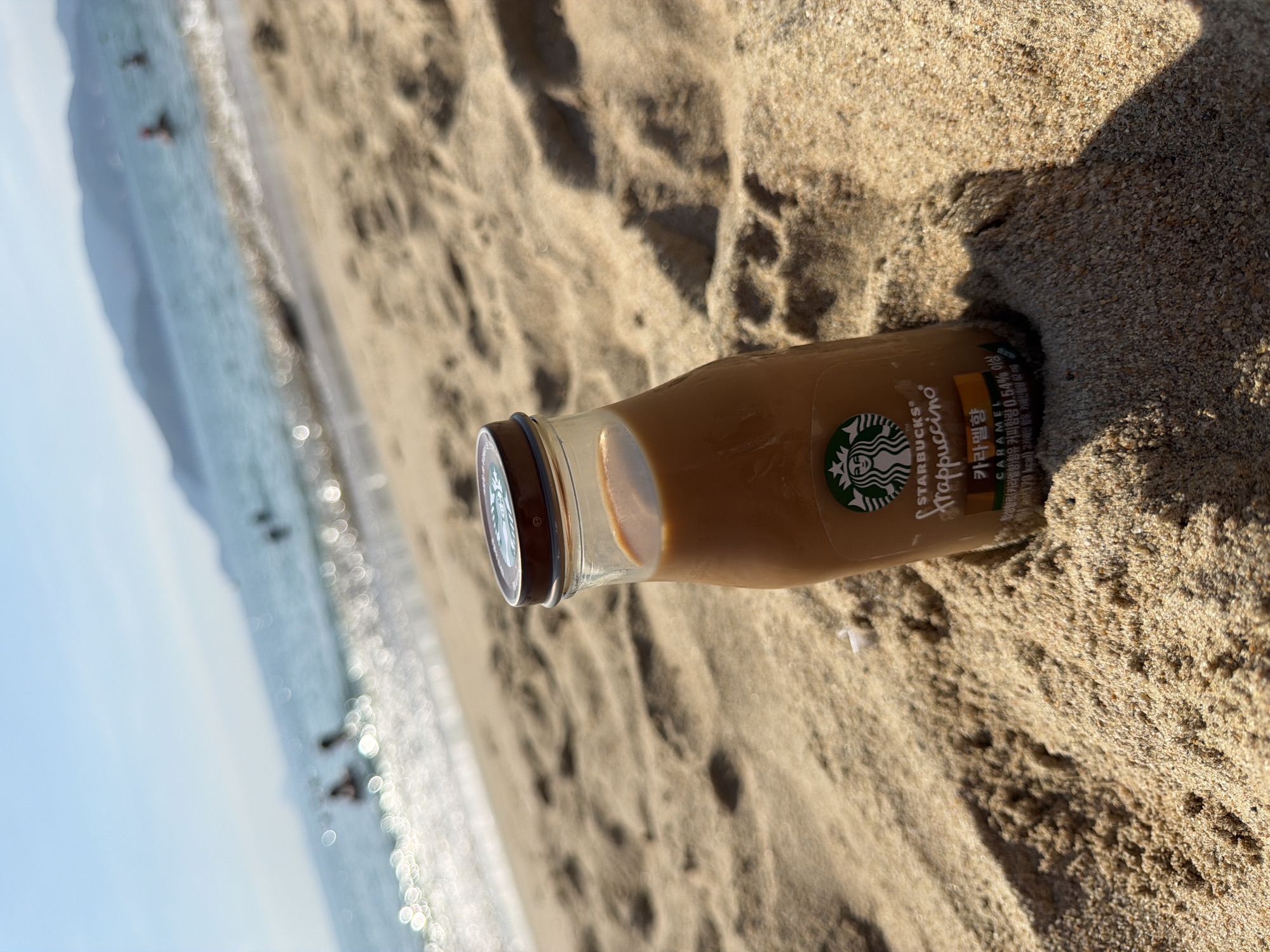 A Starbucks Frappuccino bottle, half-buried in sand on a beach, is the primary subject. The bottle is clear glass with a brown cap and label featuring the Starbucks logo and text in English and Korean, indicating "Caramel" flavor and "Frappuccino." The background shows a sunlit ocean with gentle waves and the blurry silhouettes of people swimming. In the distance, hazy mountains are visible. The sand is a light brown, showing footprints and texture. The lighting suggests a bright, sunny day. The scene depicts a relaxed, summery atmosphere. The image is taken from a low angle, close to the sand.