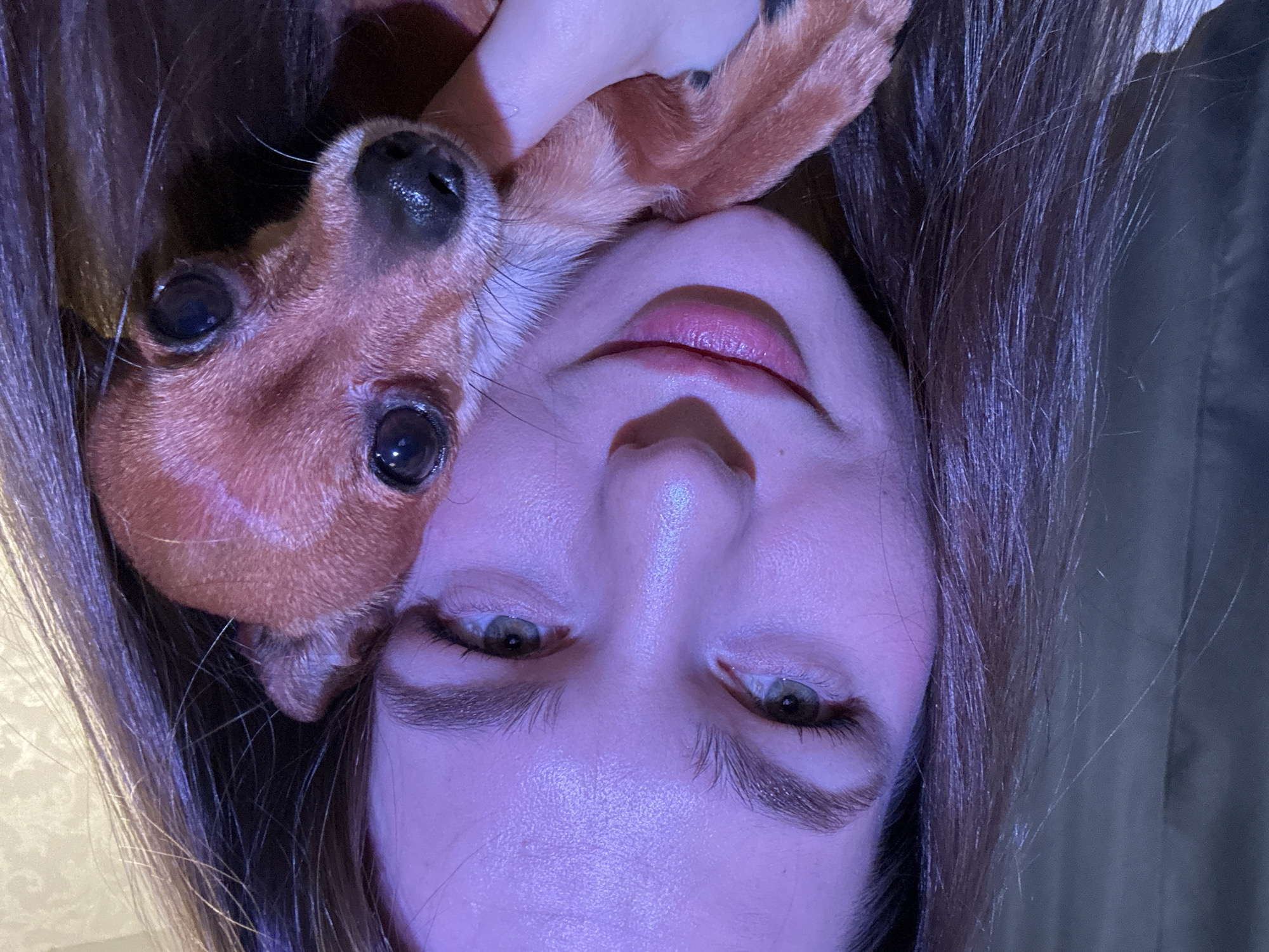 A rotated close-up photograph shows a woman's face and a small dog. The woman has fair skin, light brown hair, and green eyes. The dog, positioned above her face, is brown with dark eyes and a black nose. The scene is illuminated with a blueish light. The location is not discernible from the image.