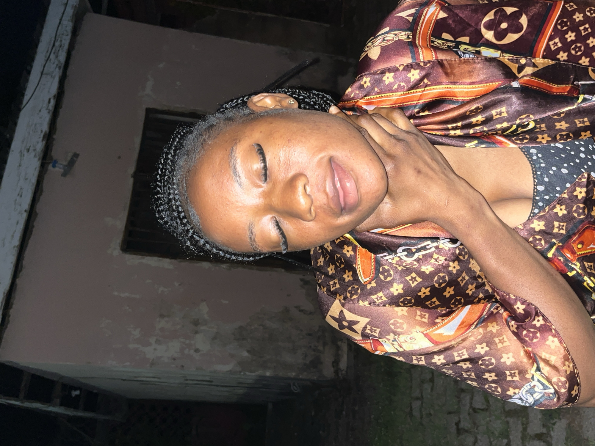 A young Black woman with braided hair and closed eyes is shown from the chest up, outdoors at night. Her right hand is clasped around her throat. She is wearing a brown satin shirt with a repeating pattern of interlocking LV symbols and flowers. Underneath, she has on a dark, polka-dotted bra. The background is a weathered, pinkish-brown building with bars on a window, and dark foliage to the left. The lighting suggests a low-light environment, possibly artificial. The overall scene is dimly lit, with the woman's face and upper body illuminated. The location appears to be an urban setting in Jos, Nigeria, given the architectural style and the woman's ethnicity.