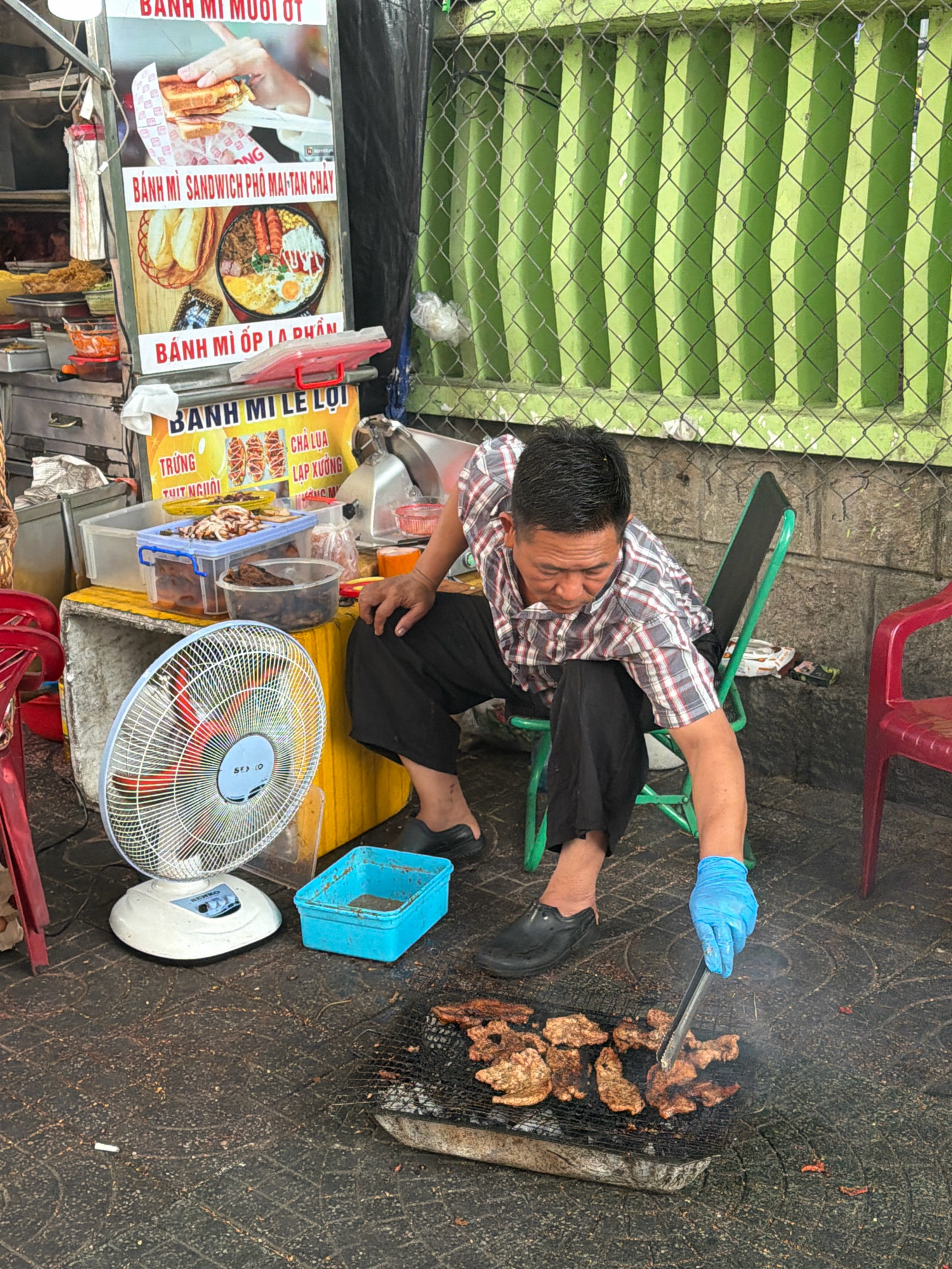 A Vietnamese man is grilling meat outdoors at a food stall in Nha Trang, Vietnam. He is wearing a plaid shirt, black pants, and blue gloves, and is using tongs to turn the meat on a charcoal grill. The grill is placed on the ground in front of him. To his left is a white electric fan. Behind him, the food stall displays menus with Vietnamese text and images of food. The background shows a green metal fence and a stone wall. The scene appears to be on a city street, likely a sidewalk, given the uneven pavement and the proximity of the stall to the street.