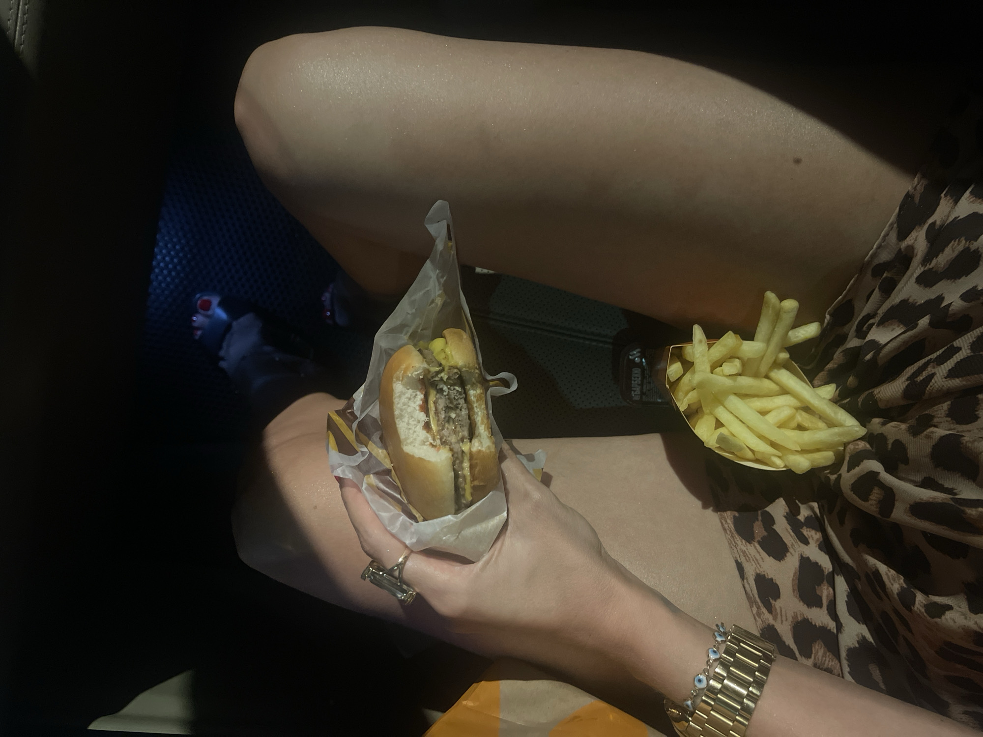 The image shows a person sitting in a car, eating a burger and fries. The person is wearing a leopard print dress and a gold bracelet. A ring is on their hand. The burger is partially eaten and wrapped in paper. The fries are in a small container. A bottle labeled “Barbecue” in Russian is partially visible. The location is likely a car in an unknown area. Without further information, the specific location in Moscow, Russia, cannot be determined.