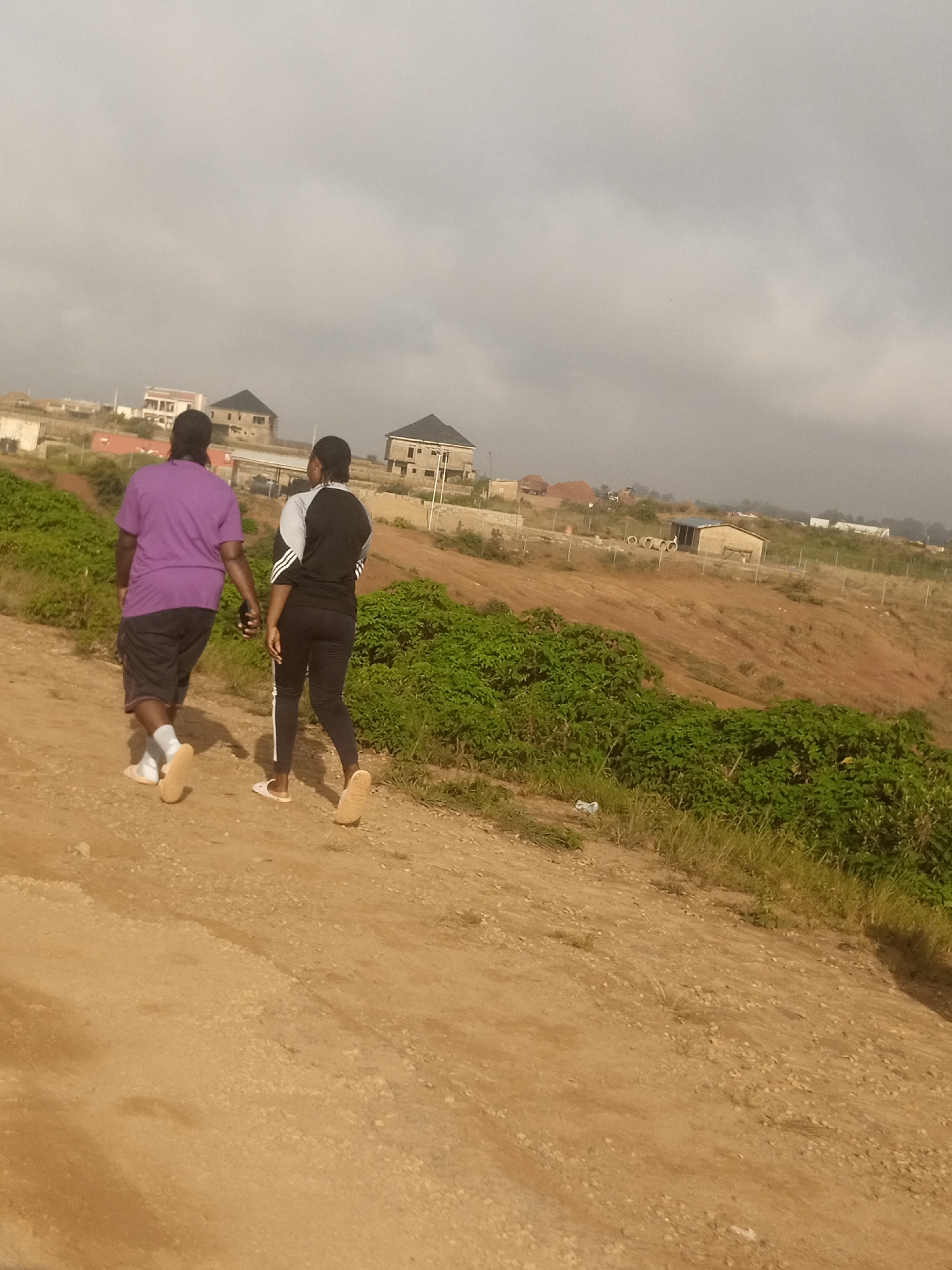 In the city of Kwang, Nigeria, two individuals, one wearing a purple t-shirt and dark shorts, and the other in a black and grey athletic top with black leggings, are walking away from the camera on a dirt path. The person in purple is holding a dark object, possibly a phone, in their right hand. They are walking side-by-side, with the person in purple slightly ahead. The ground is a dusty, sandy terrain with scattered small pebbles and dry grass. To the left and behind them, there are lush green bushes and some scattered vegetation. In the background, there are several unfinished or newly constructed buildings with various architectural styles, suggesting an ongoing development or a residential area. The sky is overcast with a hazy, greyish appearance, and the overall lighting indicates either early morning or late afternoon. The environment appears to be an outdoor, semi-urban or rural setting under a cloudy sky.