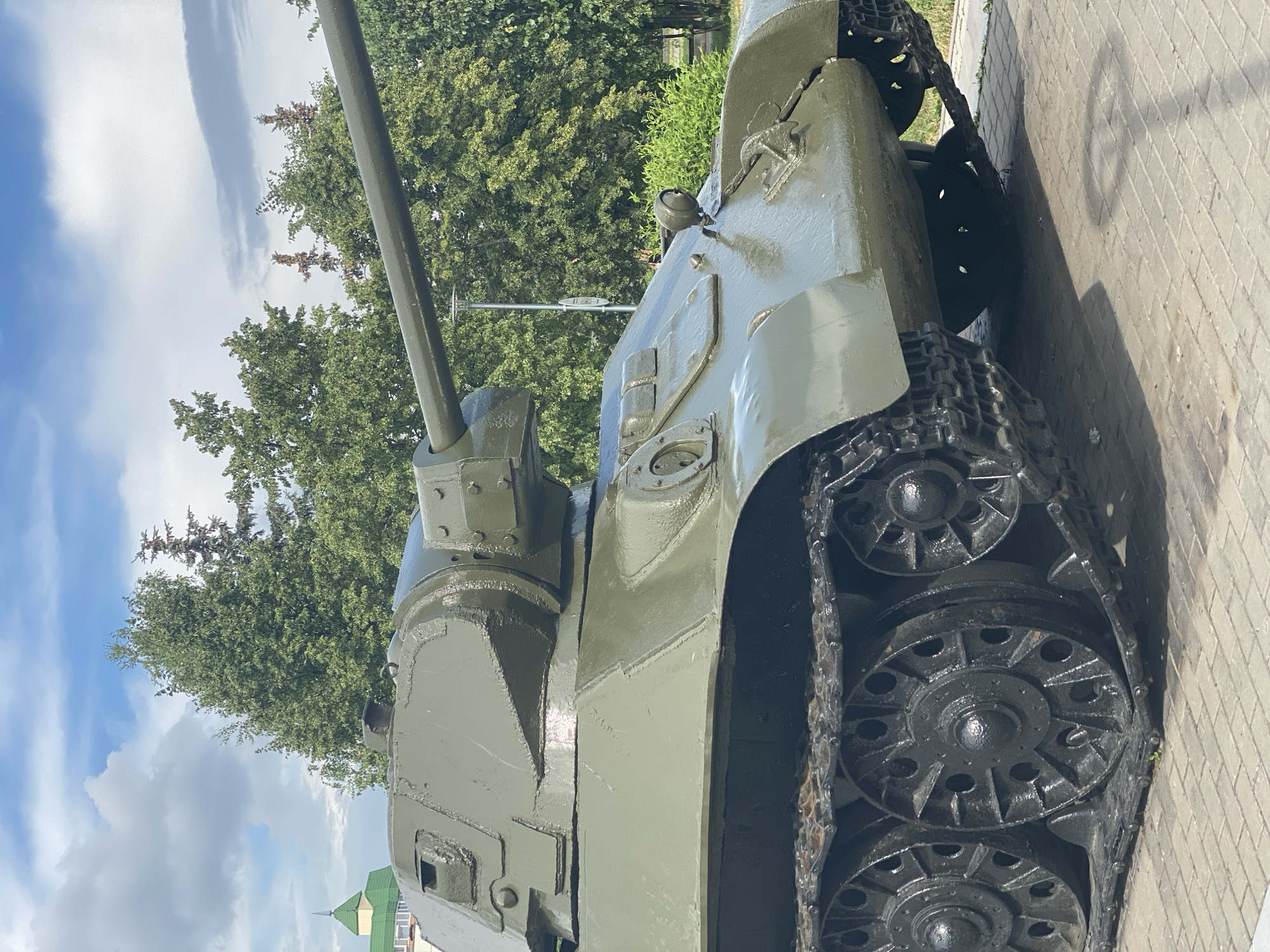 The image shows a close-up of a green military tank displayed outdoors. The tank is positioned at an angle, with its turret and gun barrel prominently visible. Its tracks and road wheels are also visible, resting on a paved surface. In the background, there are trees and a cloudy sky. The tank is a static display, likely a monument or part of an exhibit. There are no people or actions depicted in the image. The specific location in Novokuznetsk, Russia, is not identifiable from the image alone.