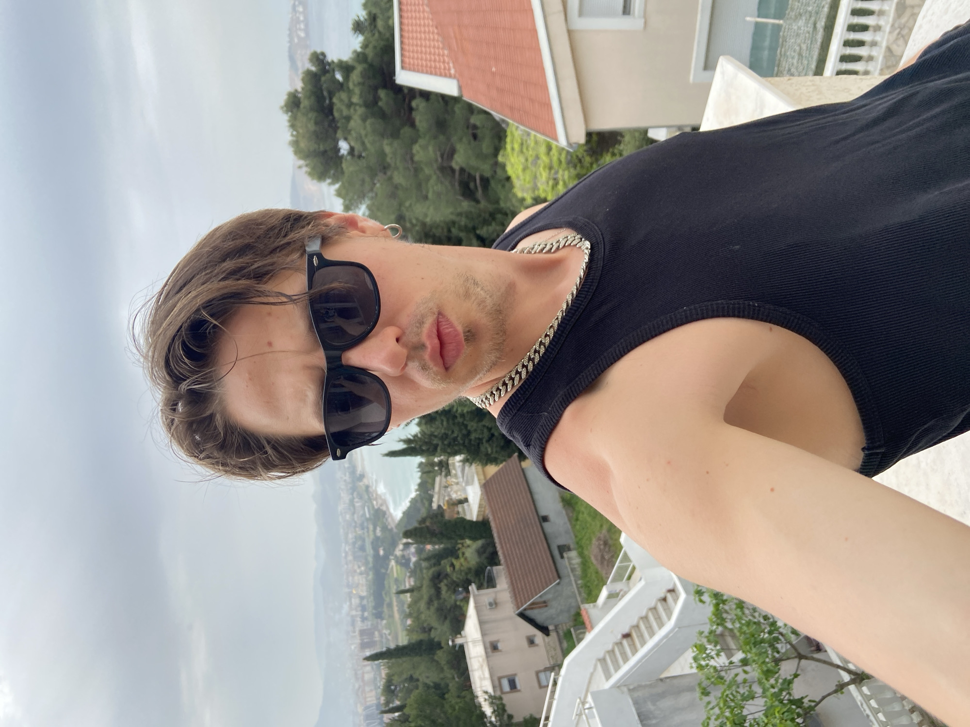 A pale-skinned man with sunglasses and a black tank top is the subject of this selfie, likely taken on a balcony. He has short brown hair, a silver chain necklace, and a small hoop earring in his left ear. In the background, the cityscape of Šušanj, Montenegro, is visible. The buildings are primarily white with red tile roofs, interspersed with green trees and vegetation. The sky is overcast. A section of a white staircase is visible on the right side of the frame.