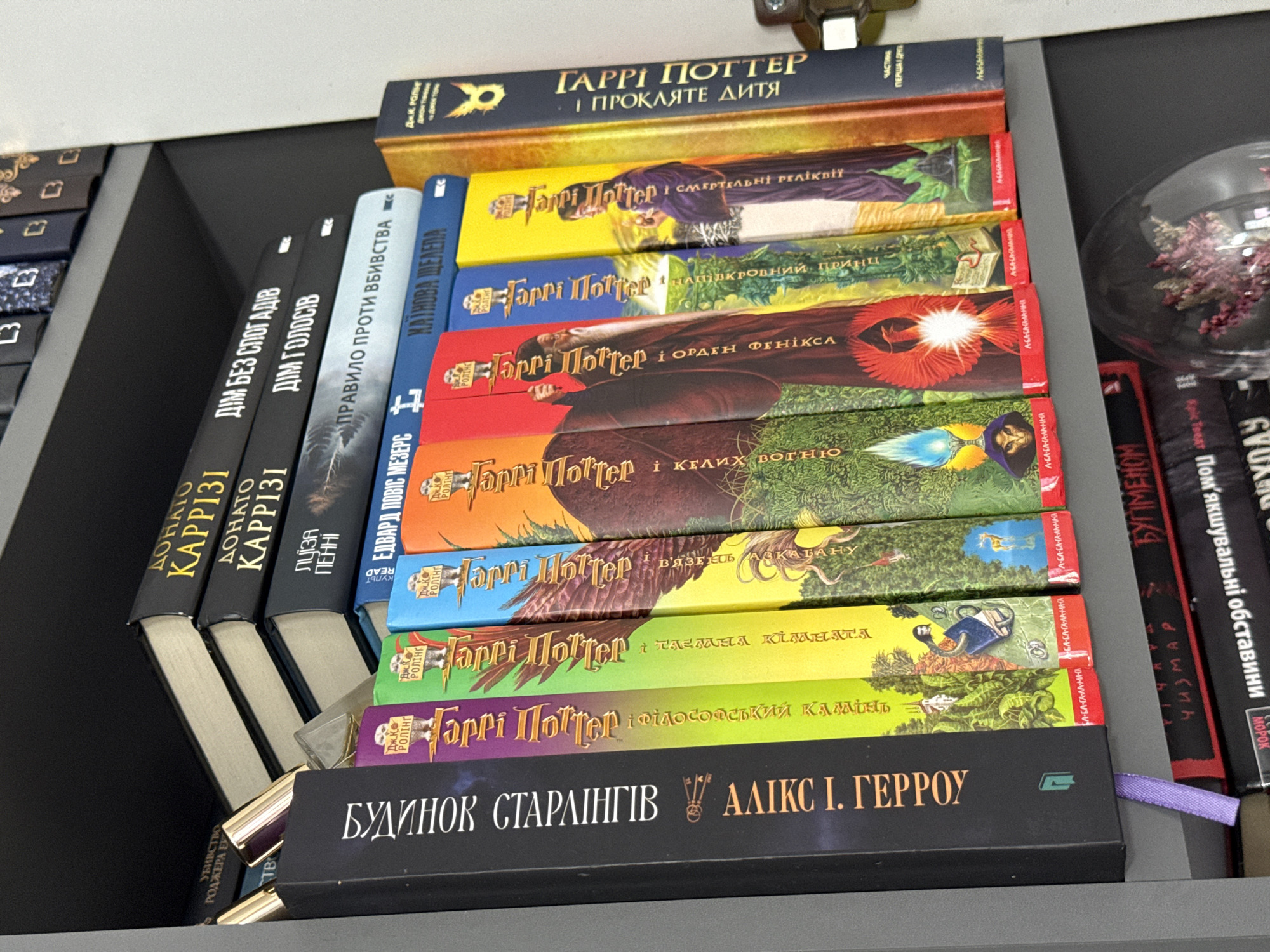 The image shows a shelf of books, primarily the Harry Potter series written in Ukrainian. The books are arranged vertically on a shelf, with the spines facing outwards. The titles are in Ukrainian, with "Гаррі Поттер" (Harry Potter) appearing prominently. Some of the visible titles include "І ПРОКЛЯТЕ ДИТЯ" (The Cursed Child), "І СМЕРТЕЛЬНІ РЕЛІКВІЇ" (Deathly Hallows), "НАПІВКРОВНИЙ ПРИНЦ" (Half-Blood Prince), "І ОРДЕН ФЕНІКСА" (Order of the Phoenix), "І КЕЛИХ ВОГНЮ" (Goblet of Fire), "ТАЄМНА КІМНАТА" (Chamber of Secrets), and "ФІЛОСОФСЬКИЙ КАМІНЬ" (Philosopher's Stone).
In addition to the Harry Potter books, there are other books on the shelf. One book on the top shelf has a dark blue cover with gold lettering that reads "ГАРРІ ПОТТЕР І ПРОКЛЯТЕ ДИТЯ". There is also a book with the title "БУДИНОК СТАРЛІНГІВ" (House of Whispers) and "АЛІКС І. ГЕРРОУ" (Alex I. Harrow) written on it.
To the right of the books, a decorative glass orb containing dried flowers is visible, hinting at an indoor setting, possibly a home or a bookstore. The overall lighting suggests it is daytime, but no specific time of day can be determined. The image is taken from a slightly elevated angle, looking down at the books. The shelf appears to be made of gray material. The context of Łódź, Poland, is provided but not visually evident in the image itself.