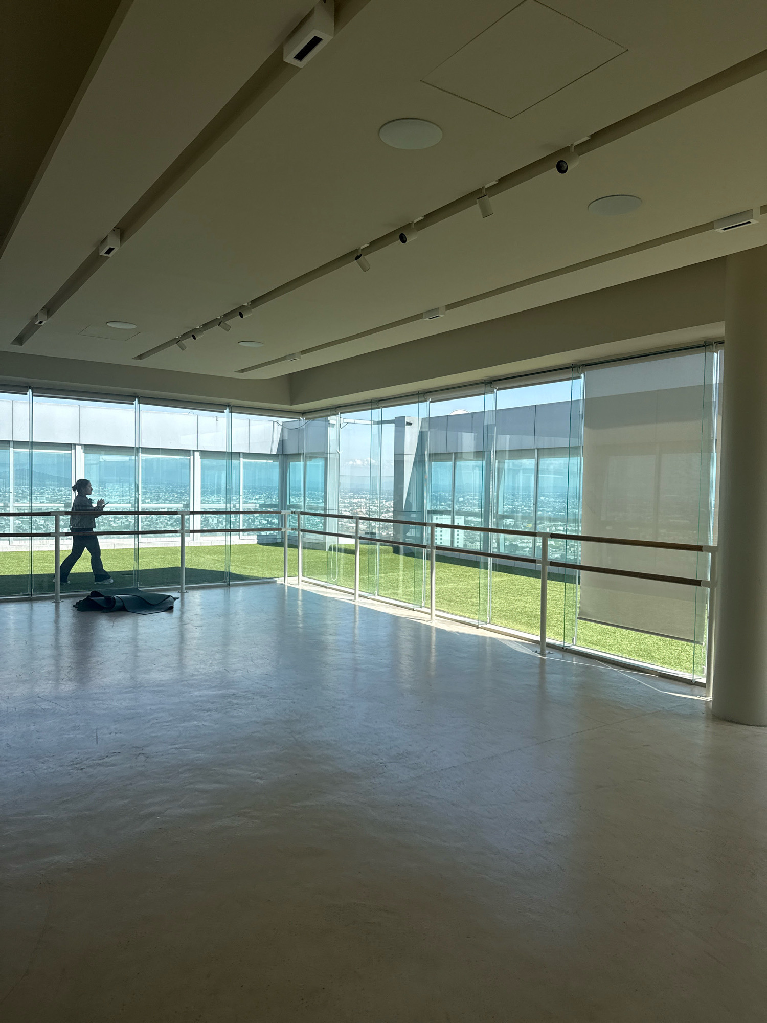 A person is walking indoors across a polished concrete floor towards a wall of floor-to-ceiling windows that overlook a city. The person is wearing dark athletic pants and a light-colored long-sleeved shirt, and appears to be holding a dark object in their right hand. A black yoga mat is lying on the floor near the person. The room has a high ceiling with recessed lighting and security cameras. A ballet barre is positioned in front of the windows, separating the interior space from an outdoor area with artificial turf. The city below is a vast expanse of buildings and infrastructure, suggesting a high-rise location in an urban environment. The scene is captured in daylight, with natural light streaming through the windows and casting shadows on the floor. The location is identified as San Bernardino Tlaxcalancingo, Mexico.
