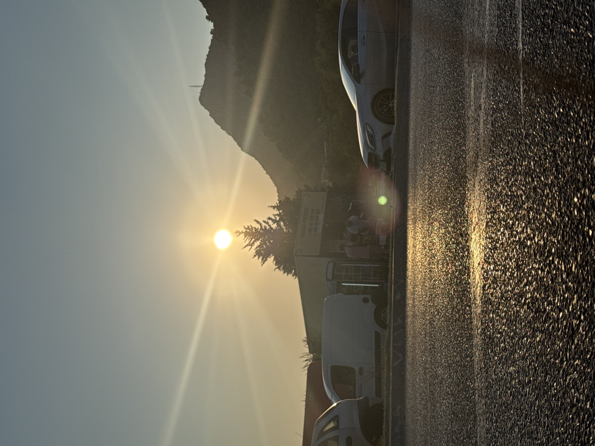 The image captures a sunset scene on a road in Sutomore, Montenegro. The sun, a bright orb with visible rays, is low in the sky, casting a warm golden light. A mountain range forms the backdrop, silhouetted against the hazy sky. In the foreground, the asphalt road is wet and reflects the sunlight, creating a shimmering effect. Several vehicles are visible, including a white van parked by the side of the road and a silver car positioned further down. A low building, possibly a shop or kiosk, is situated behind the van, with some individuals standing near it. The overall atmosphere is serene, with the setting sun illuminating the landscape.
