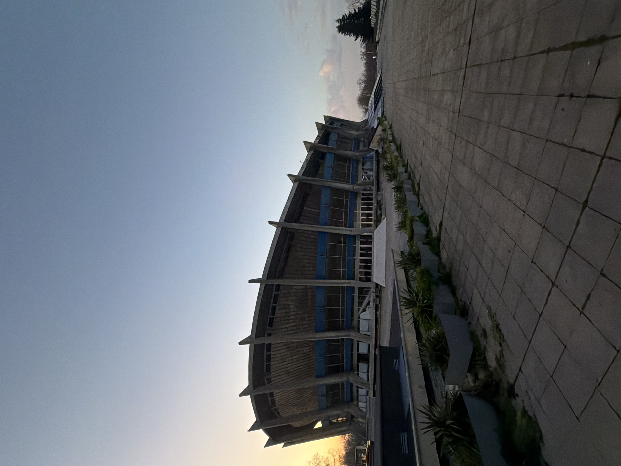 The image shows the Palace of Culture and Sports in Varna, Bulgaria. The building has a distinctive architectural design with angled supports and large windows. It is located adjacent to a paved plaza with greenery. The sky is light blue with a hint of sunrise or sunset. There are no visible people in the image.
