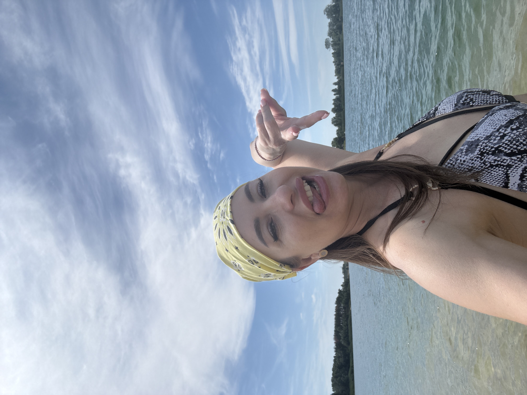 A woman wearing a yellow bandana and a snakeskin bikini poses for a selfie at the edge of a body of water. She is sticking her tongue out and making a hand gesture with her right hand. The background shows a calm body of water with a tree line in the distance under a partly cloudy sky. This is the Yelyzavetivka Reservoir in Yelyzavetivka, Ukraine.