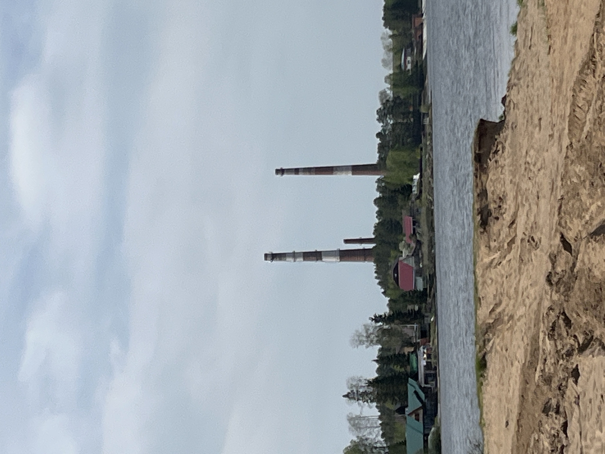 The image depicts a landscape near a body of water, possibly a river or lake, on what appears to be a cloudy day. In the background, there are tall industrial chimneys with alternating red and white bands, suggesting a power plant or similar facility. The buildings and structures along the waterfront indicate the proximity of a town or city. The foreground features a sandy or earthen embankment leading down to the water's edge.
Based on the presence of the distinctive chimneys, the location can be identified as Chaikovsky, Russia.