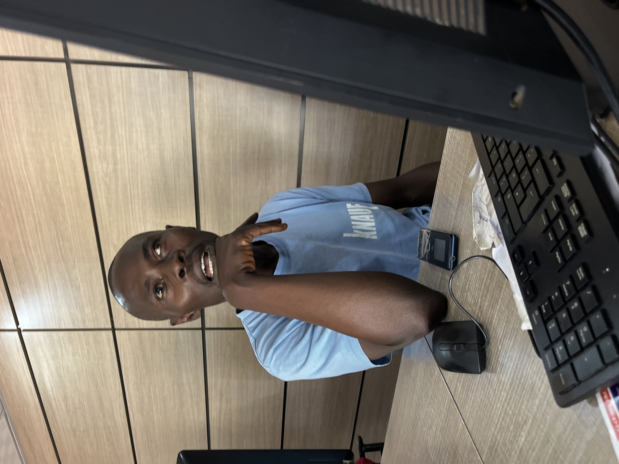 A man with a shaved head and dark skin is sitting at a desk, looking at the camera with his mouth open. He is wearing a light blue t-shirt with text that appears to read "IKNAUF". His right arm is raised, with his index finger extended, as if he is making a point. He appears to be mid-sentence, with an engaged and expressive look on his face.
The desk is made of light wood with visible grain. On the desk, there is a black computer keyboard, a black computer mouse, and a small black electronic device with a blue screen. A piece of paper is visible near the keyboard. The background consists of vertical wooden panels. The lighting suggests an indoor setting, possibly an office or workspace, during the daytime. The overall impression is that the man is actively engaged in a conversation or presentation.