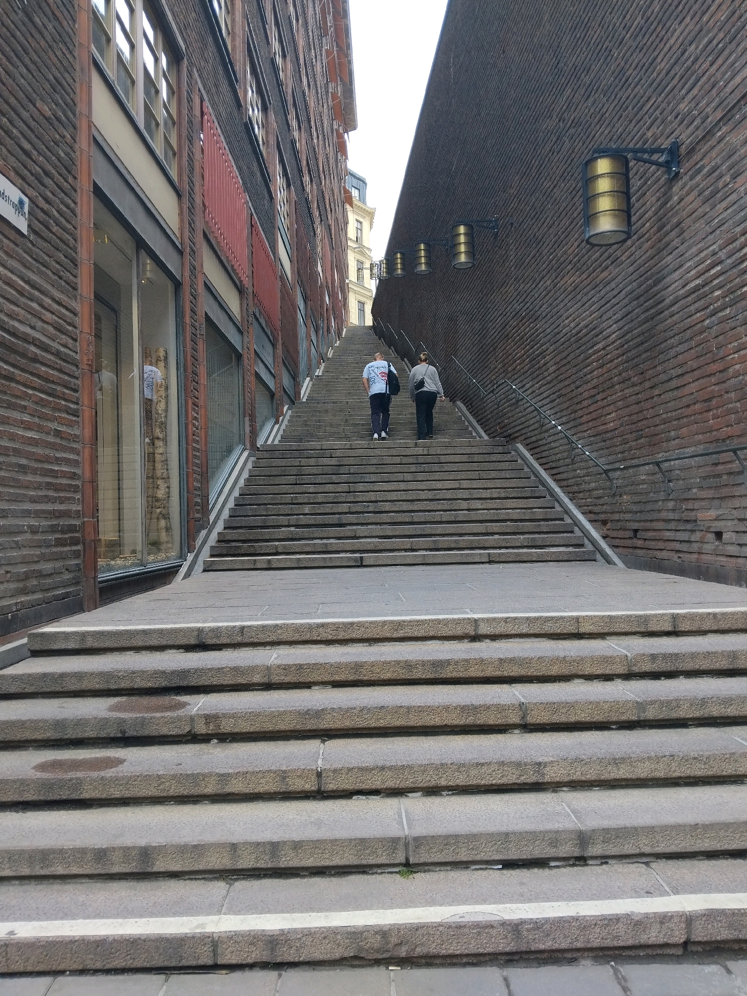 This image depicts an outdoor view of a wide, outdoor stone staircase in Stockholm, Sweden. The staircase is flanked by two buildings. The building on the left is made of dark brick and features several large windows on its lower floors, with a sign that reads "Sundströms" visible. The building on the right is also made of dark brick and has a series of cylindrical wall lamps mounted on it. Two individuals are ascending the staircase. The person in front is wearing a light-colored t-shirt and dark pants, carrying a backpack. The person behind them is wearing a gray long-sleeved shirt and dark pants, also carrying a shoulder bag. The sky above is overcast.