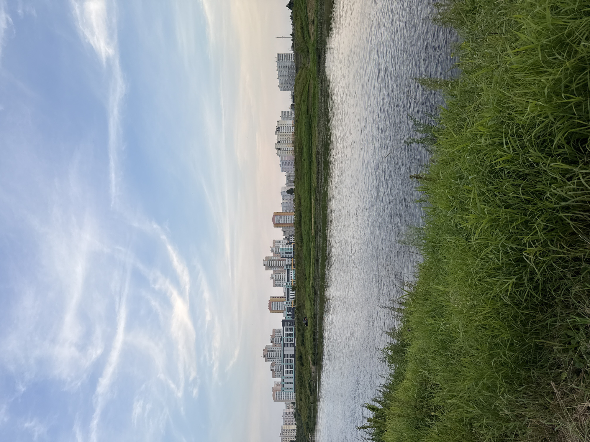 The image depicts a wide river flowing in the foreground, with lush green grass growing along its banks. In the middle ground, a grassy embankment separates the river from a line of modern apartment buildings and some smaller structures. The buildings are of varying heights and colors, with some featuring balconies and patterned facades. A few trees are scattered among the buildings. The background is dominated by a pale blue sky with wispy white clouds. The overall scene suggests an urban or suburban landscape on a clear day, with elements of nature and residential development coexisting. The context provided indicates this is a scene in Mahilyow, Belarus.