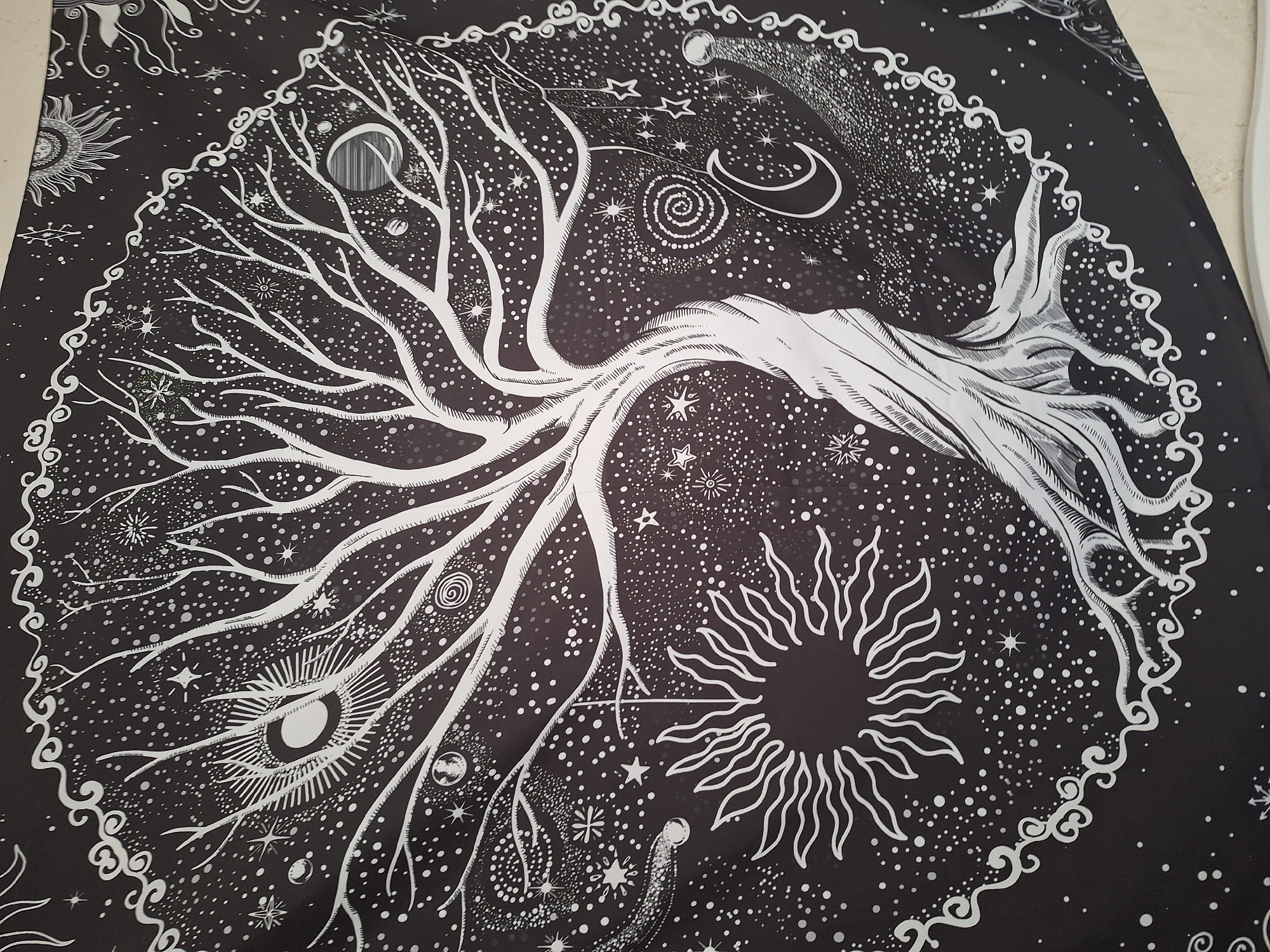 The image is a black and white tapestry featuring a stylized tree of life design. The tree's trunk and branches are white against a black background filled with various celestial elements, including stars, constellations, a crescent moon, planets, a comet, and sunbursts. The tree is centrally located and dominates the composition. The entire scene is encircled by a decorative border made of white swirls.
The image does not depict any location in the city of Panagyurishte, Bulgaria, nor any people, subjects, objects, actions, interactions or the environment pertaining to a specific place. It is a depiction of a symbolic, artistic pattern on a flat surface.