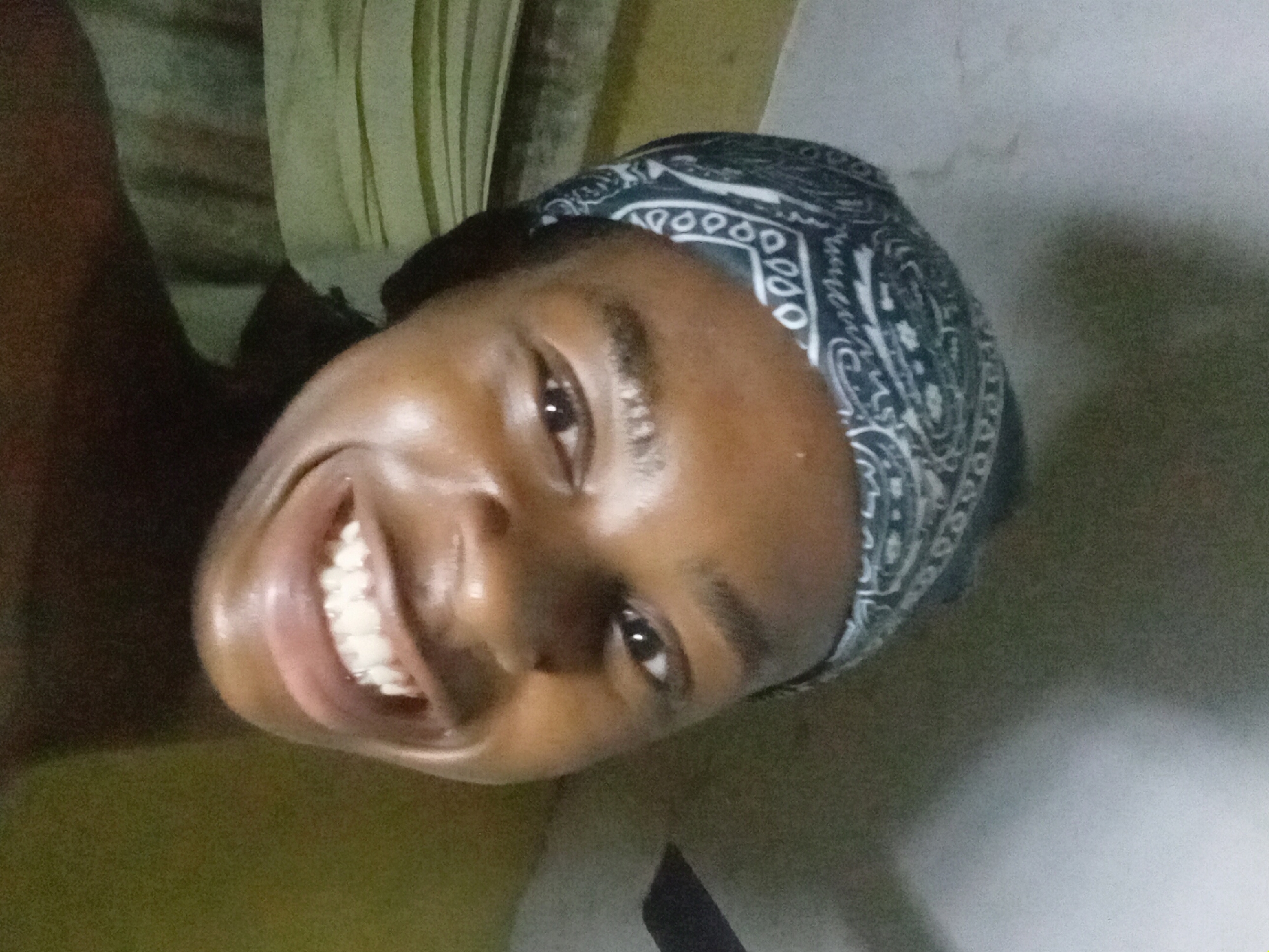 A medium close-up shot reveals a smiling person with dark skin, a visible set of white teeth, and a black and white patterned bandana covering their hair. The person is positioned at an angle, facing the camera. The location is indicated as Nkpor, Nigeria, though specific environmental details such as buildings or landmarks are not visible in the frame. A portion of a green and yellow striped object is present in the top left corner of the frame. The wall behind the person is primarily gray.