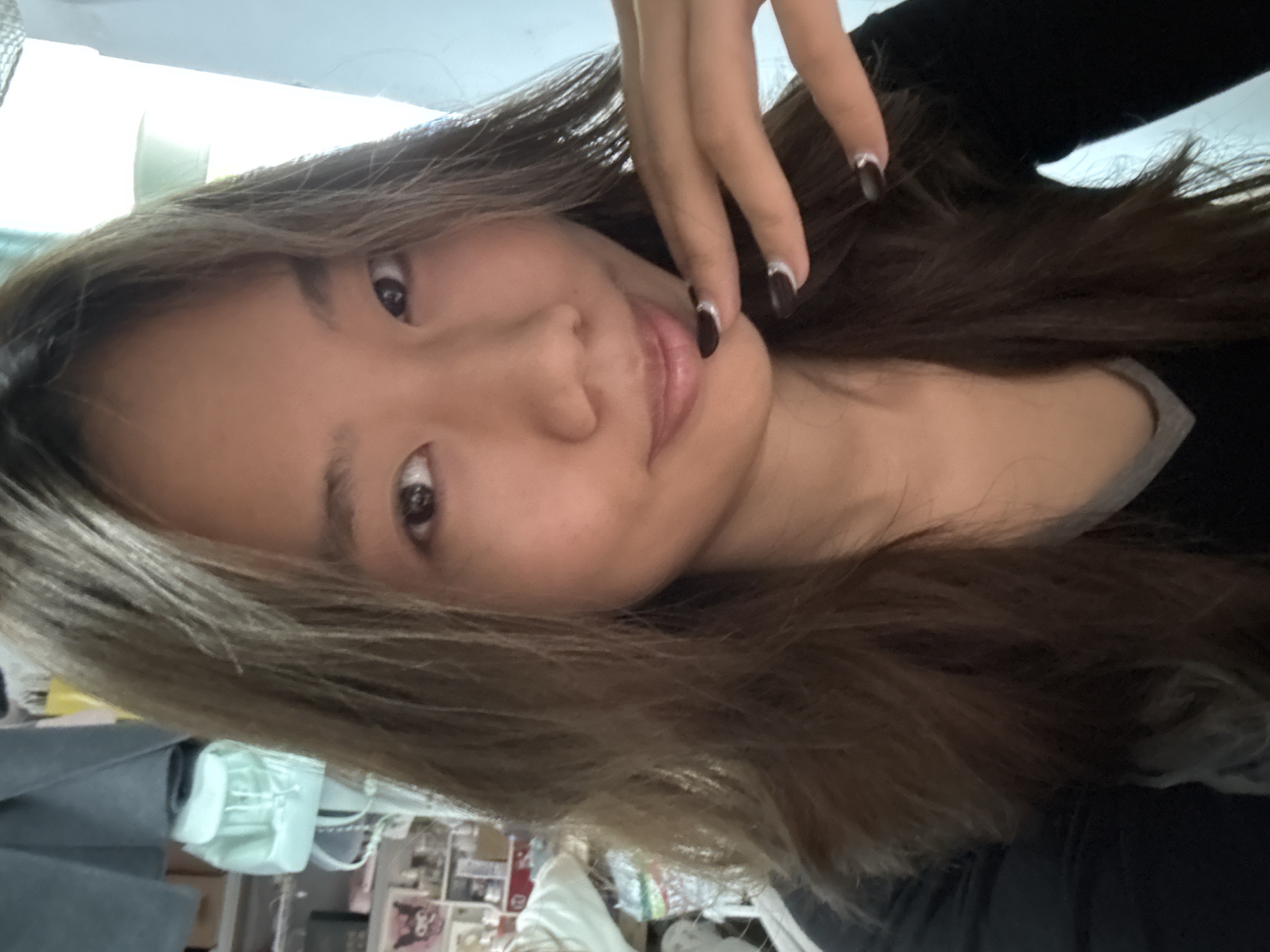 The image depicts a woman with brown hair, fair skin, and dark eyes. Her face is turned towards the camera. She is wearing a dark-colored top with a grey neckline. Her hand is near her face, showing dark-colored nails with light trim. The background shows a cluttered room with furniture, objects, and white fabric. The location is the Nan'an District, China.