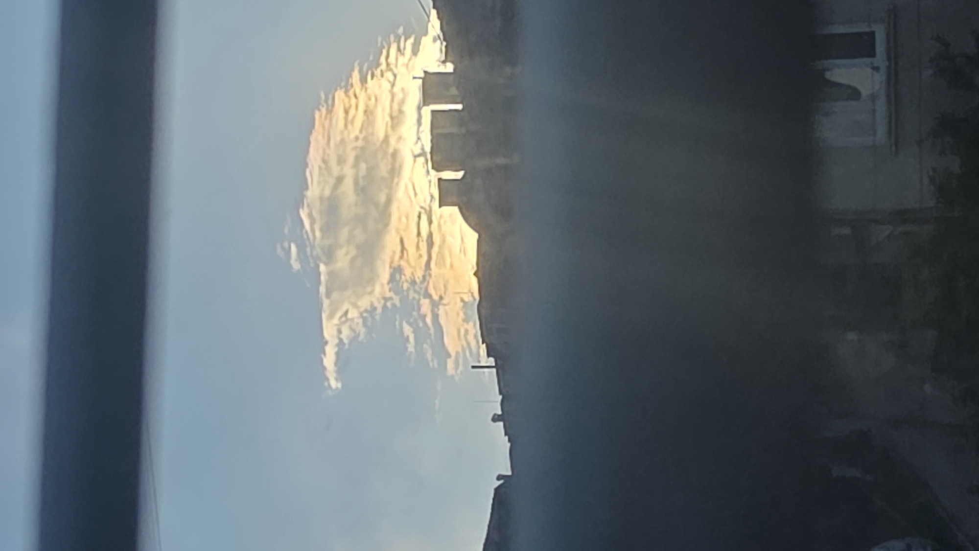 The image shows a view from a window. The sky is visible with clouds illuminated by the setting sun, casting a golden hue. In the background, a city skyline is vaguely discernible, with buildings and structures. On the right side of the frame, a window and a corner of a building with greenery are visible, suggesting a residential area. Based on the architectural style and the presence of what appears to be Cyrillic writing on a building sign (not clearly visible), it is reasonable to identify the city as Blagoevgrad, Bulgaria. The image appears to have been taken from an elevated position, likely from a high-rise building.