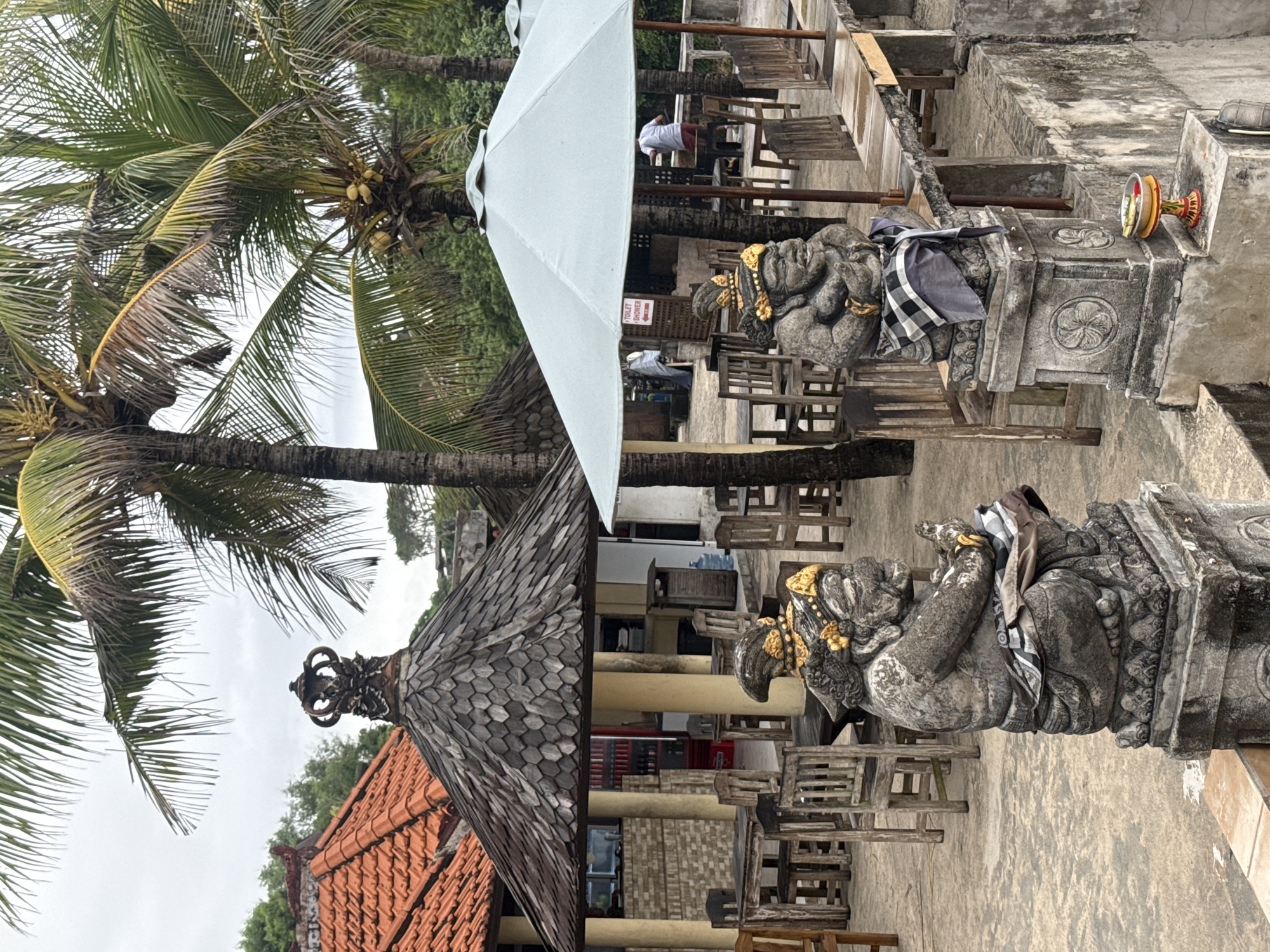 The image shows a scene in Kuta Selatan, Indonesia. Foreground elements feature two stone statues, one adorned with a black and white checkered cloth and the other decorated with gold ornaments. These statues are situated on concrete pillars.
In the background, there is an outdoor seating area with wooden chairs and tables, partially covered by a large white umbrella. Some individuals can be seen seated at the tables. A coconut tree is visible on the left side of the frame, and a building with a red-tiled roof is also present.