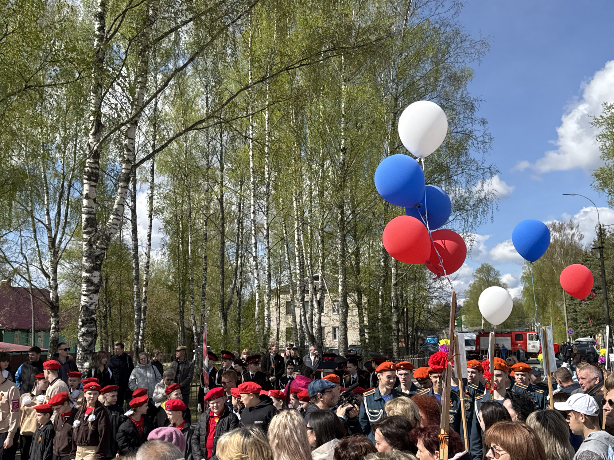 A large group of people is gathered outdoors in what appears to be a celebratory event. The location is likely Yamyshevo, Russia, although it is not directly visible in the image.
People:
The crowd is diverse, including adults and children. Many are wearing red berets and uniforms, suggesting participation in a ceremony or parade. Some individuals are in military or emergency service uniforms. There is a mix of genders and ages among the crowd.
Subjects/Objects:
Several balloons are visible in the colors of the Russian flag: white, blue, and red. There are trees in the background, many of which are birch. A fire truck is visible in the background, implying a public event or safety presence. Some participants carry wooden flagpoles, potentially for flags or banners.
Actions/Interactions:
The crowd is assembled, suggesting a gathering or procession. Some individuals appear to be looking in a specific direction, perhaps at a speaker or performance.
Environment:
The setting is outdoors on a clear day with blue skies and some clouds. The presence of trees indicates a park or green space. Buildings can be seen in the background, further suggesting a town or city environment.