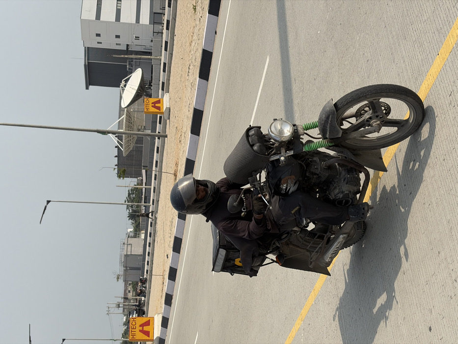 A male individual is depicted riding a dark Olink motorcycle on a paved road in Alaguntan, Nigeria. The rider wears a black full-face helmet with a dark visor, a dark long-sleeved shirt with a "Cainiao" logo, dark trousers, and dark clogs, along with dark gloves. The motorcycle features bright green front suspension components, a single round headlight, and a dark, textured cylindrical cover over the handlebars. A large black delivery box with yellow markings is affixed to the motorcycle's rear. The road surface is light grey, marked by a continuous yellow line. To the right of the road, a median with black and white striped curbing borders a sandy expanse. In the background, modern architectural structures are visible, including a white and grey building. Two large white satellite dishes are positioned on structures within the background. Yellow triangular signs with "HITECH" in black text are present in the sandy area. Tall streetlights equipped with LED fixtures line the road. A solitary figure walks in the distant background, and a dark SUV is partially visible further left. The sky is a light, hazy grey, and shadows are cast on the road surface, indicating direct sunlight.