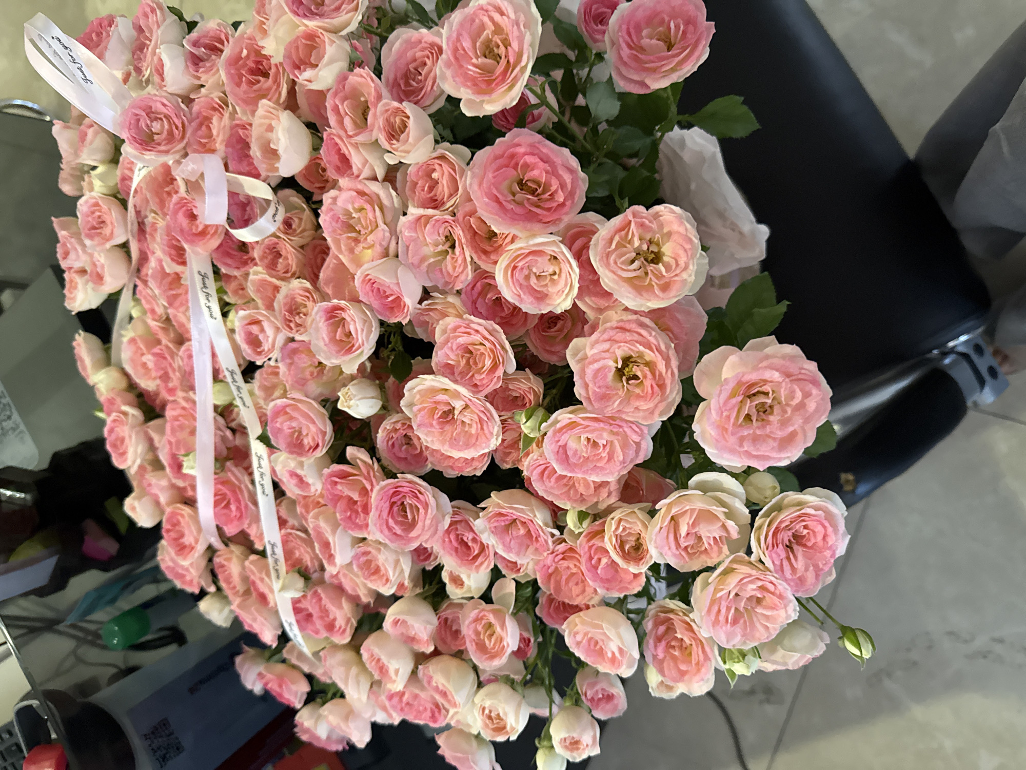 This image shows a large bouquet of light pink and white roses, possibly garden roses or spray roses, arranged in a black vase. The roses are densely packed, with many buds and fully bloomed flowers. A thin white ribbon with the text "Just for you" printed in black is draped across the bouquet. The background is blurred but appears to be indoors, with a dark surface and hints of reflective elements. Based on the information provided in the prompt, this scene is located in Bishkek City, Kyrgyzstan. There are no people visible in the image, and the focus is entirely on the floral arrangement. The overall impression is one of a carefully presented gift.