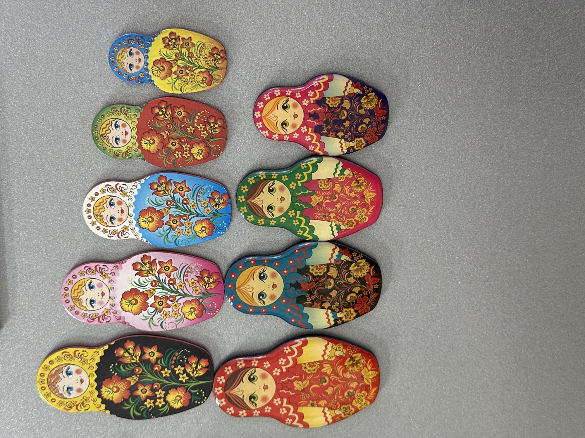 The image displays a collection of eight Matryoshka dolls arranged in two rows on a gray, textured surface. The top row features four dolls, with the largest on the left and progressively smaller dolls to the right. The bottom row contains four larger dolls, also arranged from left to right. All dolls are depicted in a frontal view and are decorated with vibrant floral patterns and facial features.
The dolls exhibit a variety of color schemes. The top row includes a black doll with yellow and red flowers, a pink doll with similar floral designs, a blue doll with red and yellow flowers, and a red doll with yellow and red flowers. The bottom row showcases a red doll with yellow accents, a blue doll with red and yellow floral patterns, a green doll with pink and yellow floral designs, and a purple doll adorned with red and yellow flowers.
The background is a plain gray surface, which appears to be a flat, horizontal plane with a subtle sparkle or texture. No specific location in Chaikovsky, Russia, can be identified from this image. The image is a still photograph of decorative objects.