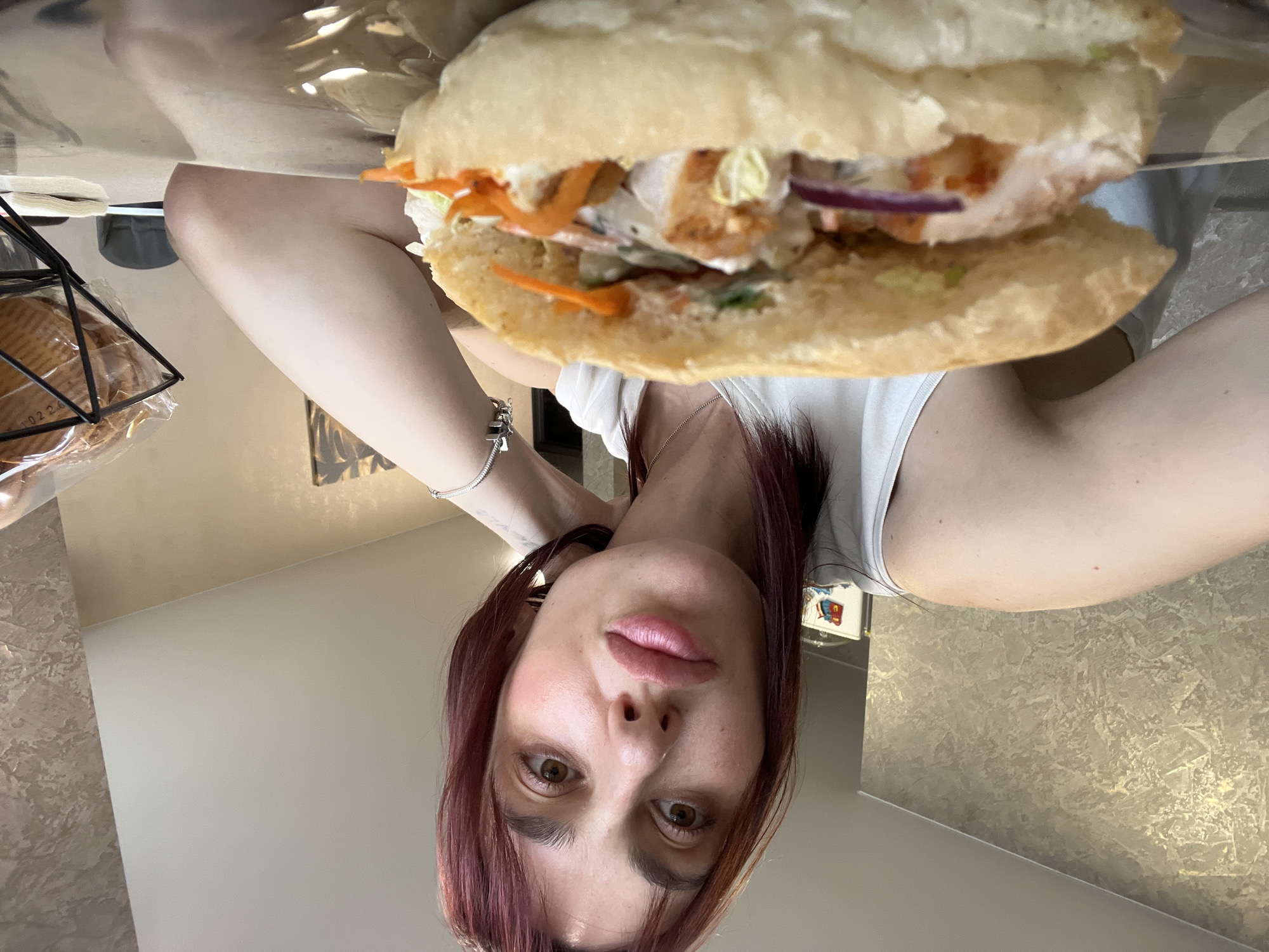 The media file shows a close-up, upside-down, selfie-style shot of a person holding a sandwich in front of their face. The person, with reddish-brown hair and light-colored eyes, is wearing a white tank top and a silver bracelet. They are looking directly at the camera with a neutral expression. The sandwich appears to be made with a long bun and filled with chicken, lettuce, shredded carrots, and red onion.
The background is blurry, but suggests an indoor setting. The lighting is soft and warm, indicating it could be daytime. The overall ambiance is casual and personal. There is no visible text in the image. The location context of Nizhny Novgorod, Russia is provided, but there are no specific visual cues within the image to confirm this location.