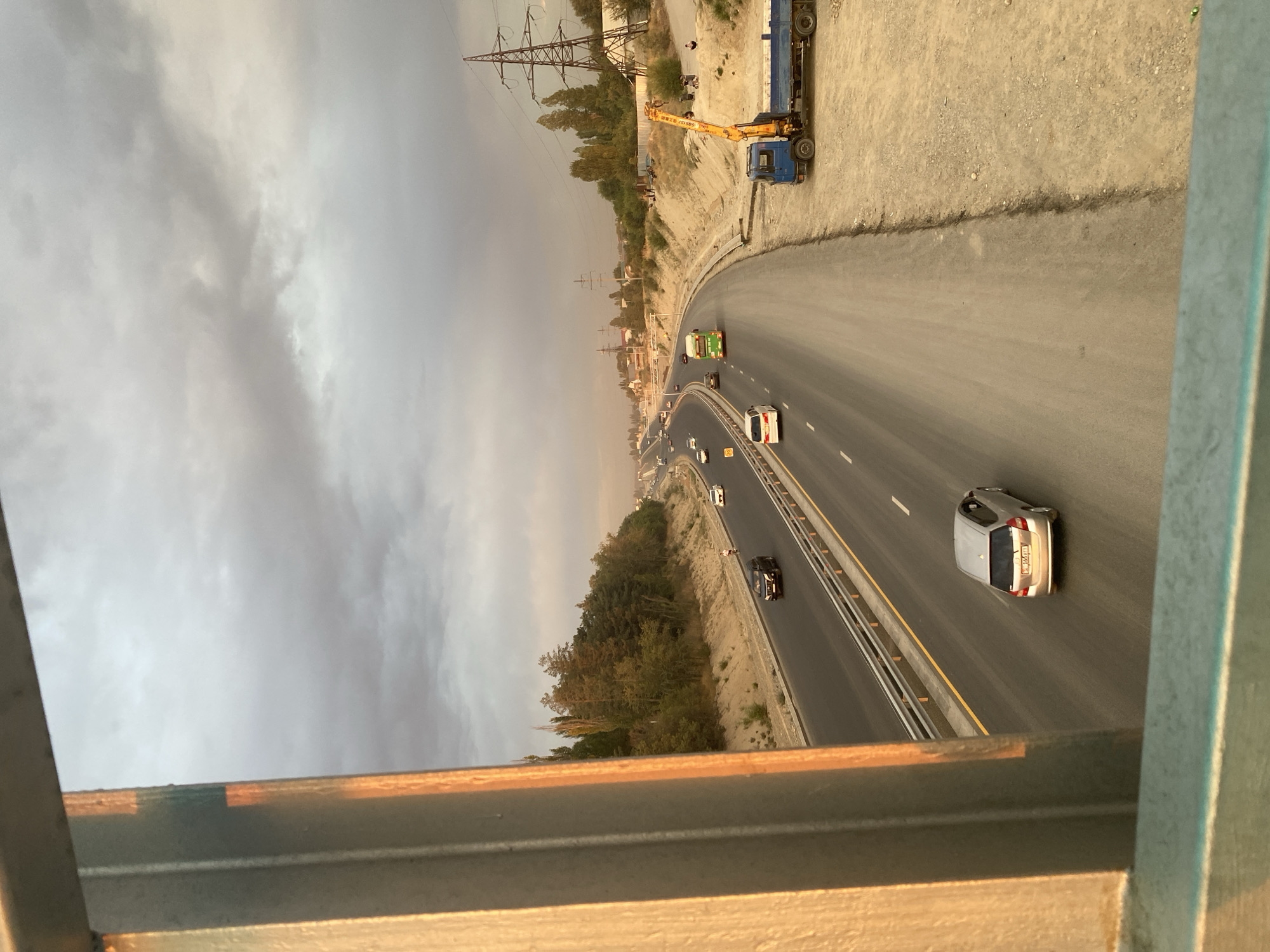 The image captures a view of a highway in Bishkek City, Kyrgyzstan, during a cloudy day with golden hour lighting. The foreground is dominated by a metal railing, likely part of a bridge or overpass, casting an orange reflection from the setting sun. The highway stretches into the distance, with multiple lanes of traffic. Cars, including a silver hatchback, a black SUV, and a white sedan, are visible on the road. A green bus is also traveling on the highway.
On the right side of the highway, there is a construction area with a blue truck equipped with a crane. The ground is dirt and gravel, with some piles of material. In the background, there are power lines with tall pylons and scattered buildings and trees. A small group of people can be seen near the construction area and on the side of the road. The sky is filled with dramatic, grey clouds, suggesting either an approaching storm or the end of a rainy day. The overall scene depicts a typical urban landscape with ongoing infrastructure development and the flow of daily life on a major road.