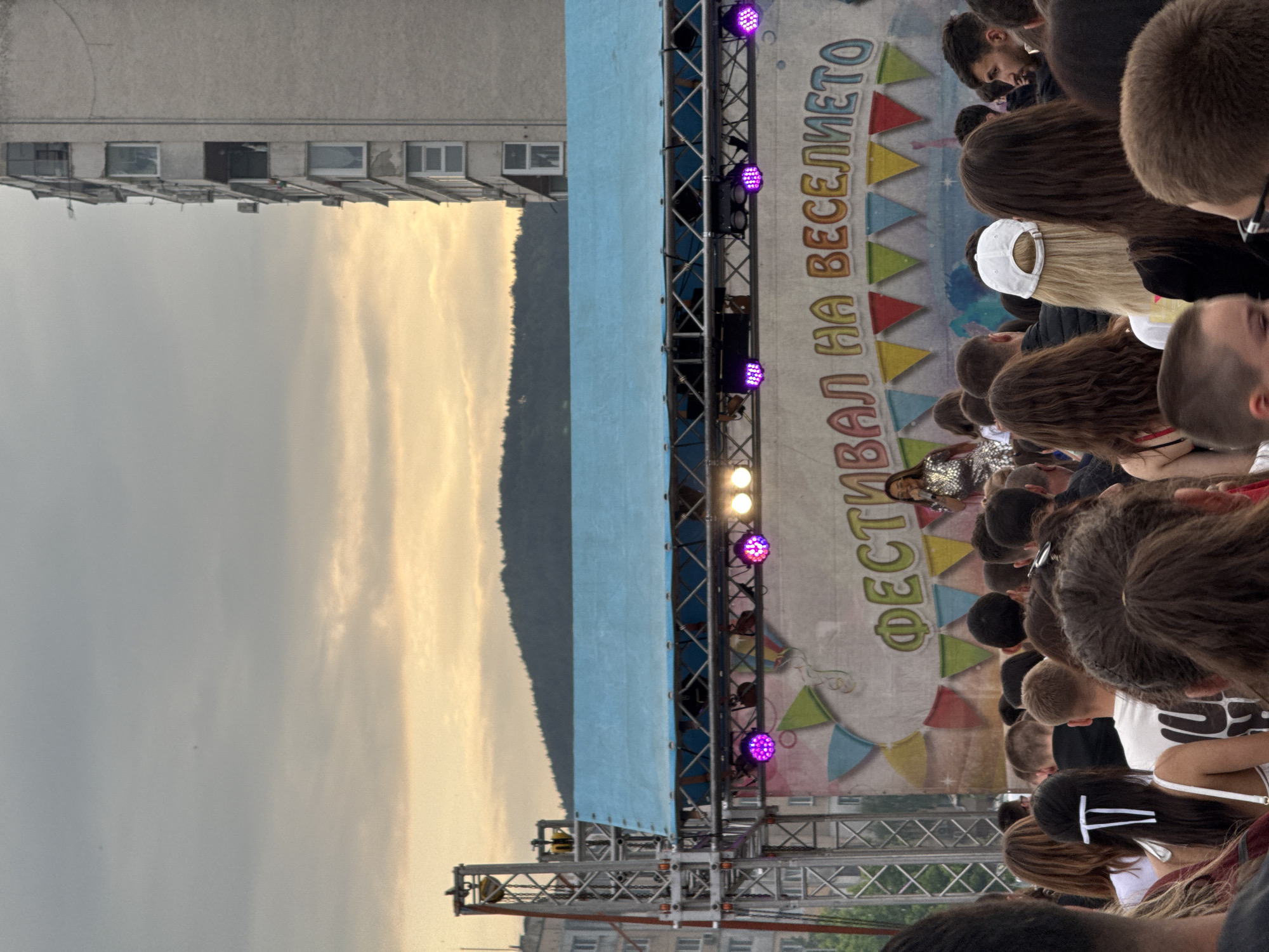 The image depicts an outdoor festival or concert scene, likely during the late afternoon or early evening. A large crowd of people is gathered in the foreground and midground, mostly viewed from behind, intently facing a performance stage positioned on the right side of the frame. One performer, visible on stage in a sequined top, is illuminated by various stage lights, including purple and white fixtures mounted on metal trussing.
The stage backdrop is a large banner featuring the Cyrillic text "ФЕСТИВАЛ НА ВЕСЕЛИЕТО," which translates to "FESTIVAL OF MERRIMENT" or "FESTIVAL OF FUN." Below the text, colorful triangular pennants decorate the banner. In the background, on the left, stands a tall, multi-story building with several windows. Beyond the urban structure, a silhouette of mountains or hills stretches across the horizon. The sky exhibits warm, golden light towards the right, transitioning to a softer grey on the left, suggesting the golden hour or an approaching sunset. The setting is identified as Shumen, Bulgaria, consistent with the visible Cyrillic text. The scene conveys a lively public gathering enjoying an outdoor performance.