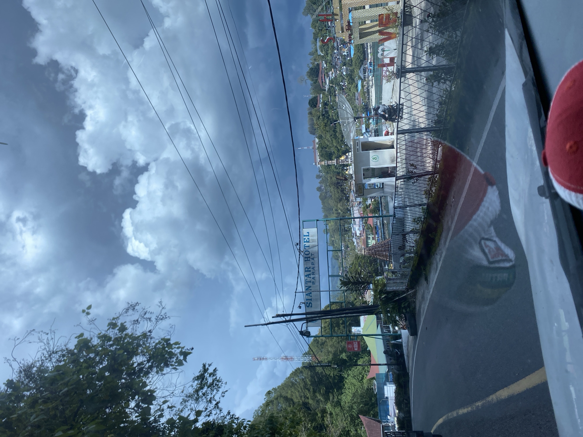 The image depicts a street scene in Tiga Raja, Indonesia, on a cloudy day. A road curves to the left, with a yellow line marking the center. On the right side of the road, there is a fence bordering an area with decorative "LOVE" letters, a small building with a satellite dish, and a motorcycle parked. In the background, buildings and vegetation cover a hillside, and a prominent "SIANTAR HOTEL PARAPAT" sign is visible. A tall red and white radio tower stands on a distant hill. The sky is filled with dark, dramatic clouds, suggesting impending rain. The reflection of the vehicle's interior, including a red and white baseball cap, is visible on the road surface.