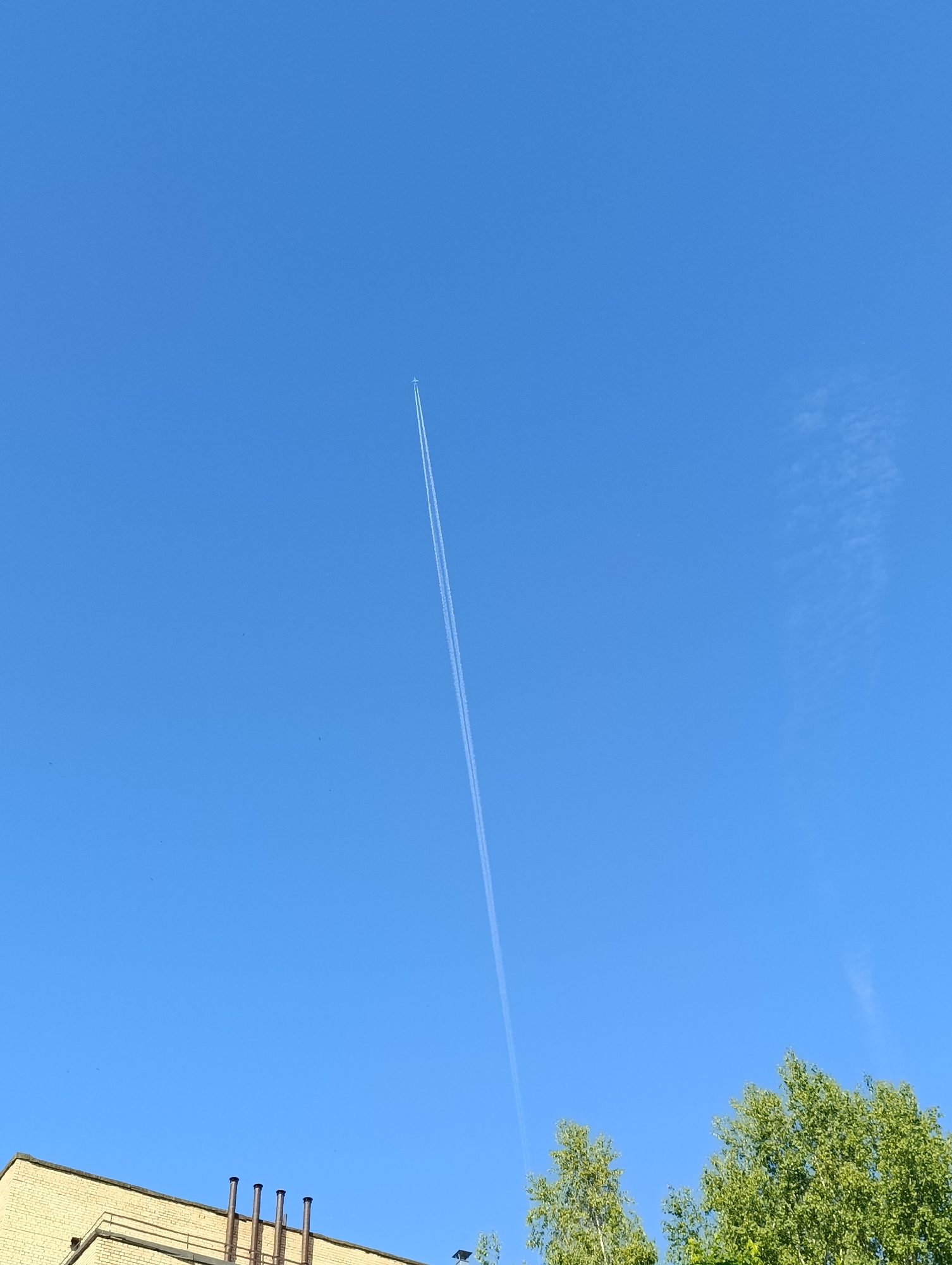 The image displays a clear, bright blue sky on a sunny day. A small, white airplane is visible high in the sky, positioned in the upper center, leaving a long, distinct white contrail that stretches diagonally downwards and to the left. The contrail appears to be initially composed of two parallel lines.
In the bottom left corner, the top section of a light-colored brick building, possibly yellow or beige, is partially visible. Several dark, cylindrical pipes, likely chimneys or ventilation shafts, protrude from the roof of this building. The bottom right corner of the frame is filled with the vibrant green foliage of several leafy trees.
The scene depicts an outdoor environment, likely urban or suburban, under a vast, open sky. The bright lighting suggests it is daytime, possibly mid-morning or afternoon, with no significant clouds other than the airplane's contrail. There are no people visible, nor is there any discernible text.