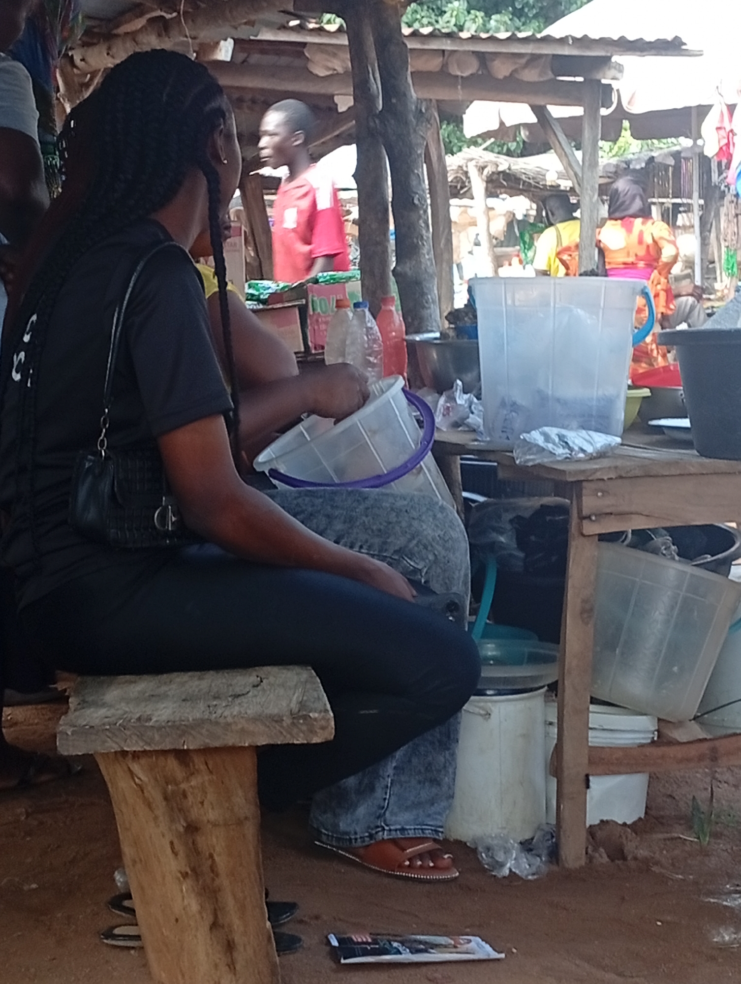 This image depicts an outdoor scene at a market stall in Andaha, Nigeria. In the foreground, a person with braided hair is seated on a wooden stool, facing away from the camera. They are wearing a black t-shirt and dark pants, with a small black purse hanging from their shoulder. To the left of this person, another individual is partially visible. In the background, several other people are present at the market. One person, wearing a red shirt, is standing. Others are seen behind a stall, where various items are displayed, including large plastic containers, buckets, bottles, and what appear to be food items or merchandise. The stall itself is constructed with wooden poles and a corrugated metal roof, typical of market structures. The ground is earthen, and there is sparse vegetation visible. The overall atmosphere suggests a busy marketplace environment.