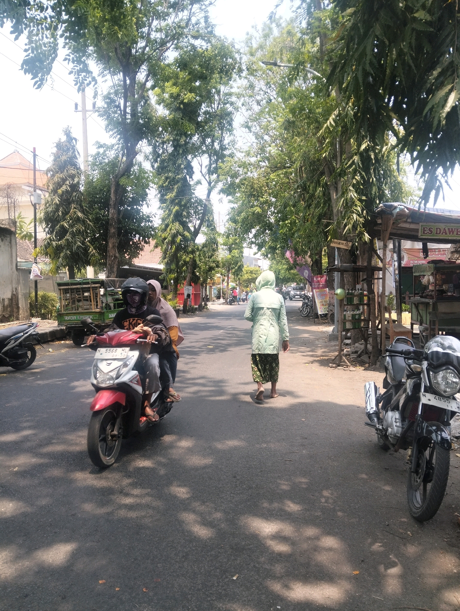 The image depicts a street scene in Semampir, Indonesia, likely on a sunny day given the shadows cast on the asphalt. In the foreground, a red and white scooter is being ridden by two individuals. The driver, wearing a black helmet and a patterned face covering, is operating the vehicle, while a passenger wearing a light brown headscarf sits behind. A black motorcycle is parked on the right side of the road. Further down the street, a person in a light green hooded jacket and patterned skirt walks away from the camera, barefoot. Various trees line the sides of the road, providing shade. A small food stall with the signage "ES DAWE" is visible on the right. In the background, a green vehicle, possibly a small truck, is parked near a building with a red roof. The road appears to be an urban or semi-urban street with scattered buildings and vegetation.