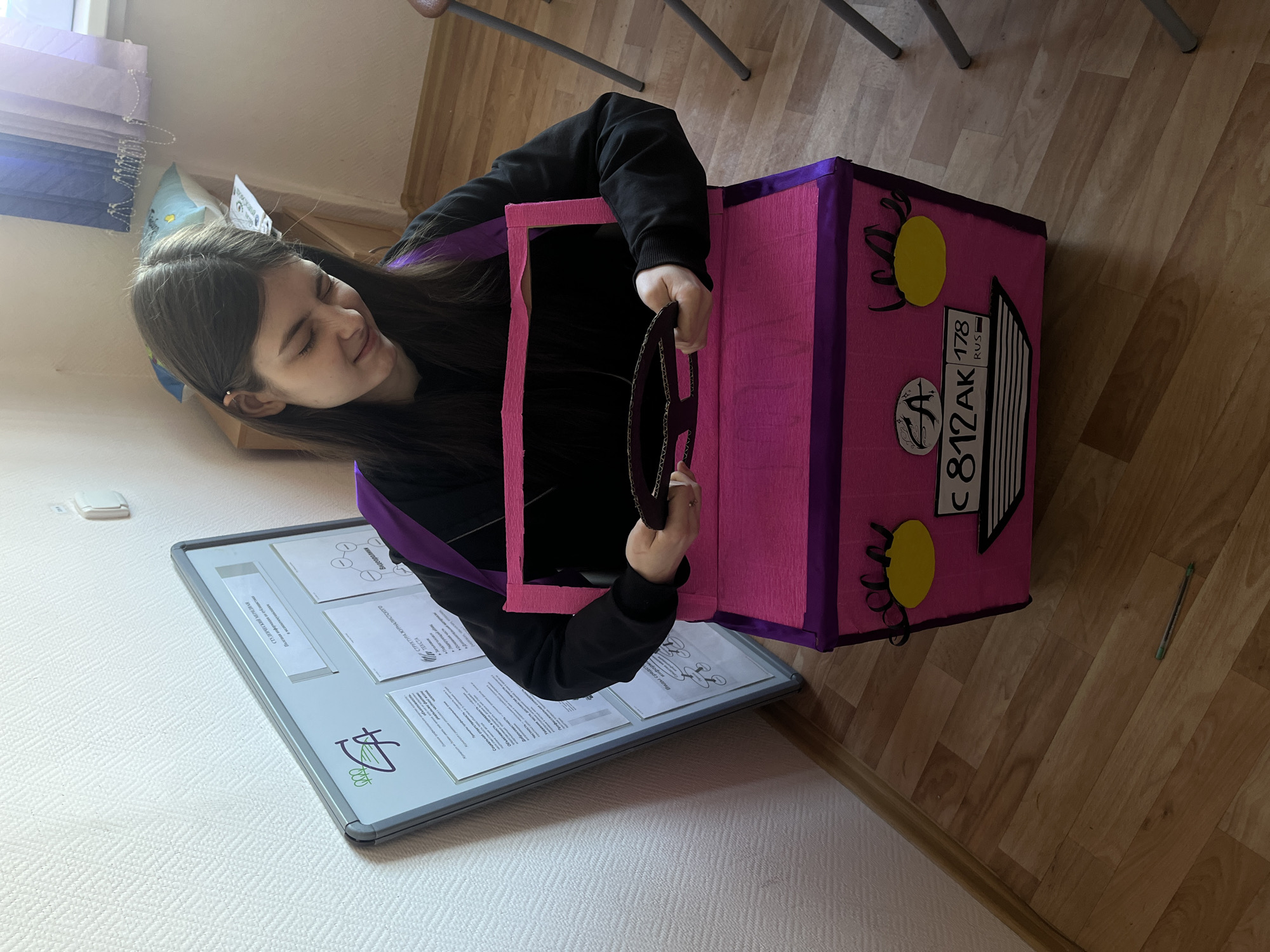 A young woman with long, dark hair is sitting inside a pink and purple cardboard box resembling a car. She is smiling and holding a cardboard steering wheel. The "car" has a license plate that reads "C 812 AK 178 RUS." The "178 RUS" indicates that the car is registered in Saint Petersburg, Russia. The scene appears to be indoors, possibly in a classroom or office, given the presence of a bulletin board with papers on the left and chairs in the background. The floor is made of wooden planks. There's a green pen near the wall.
