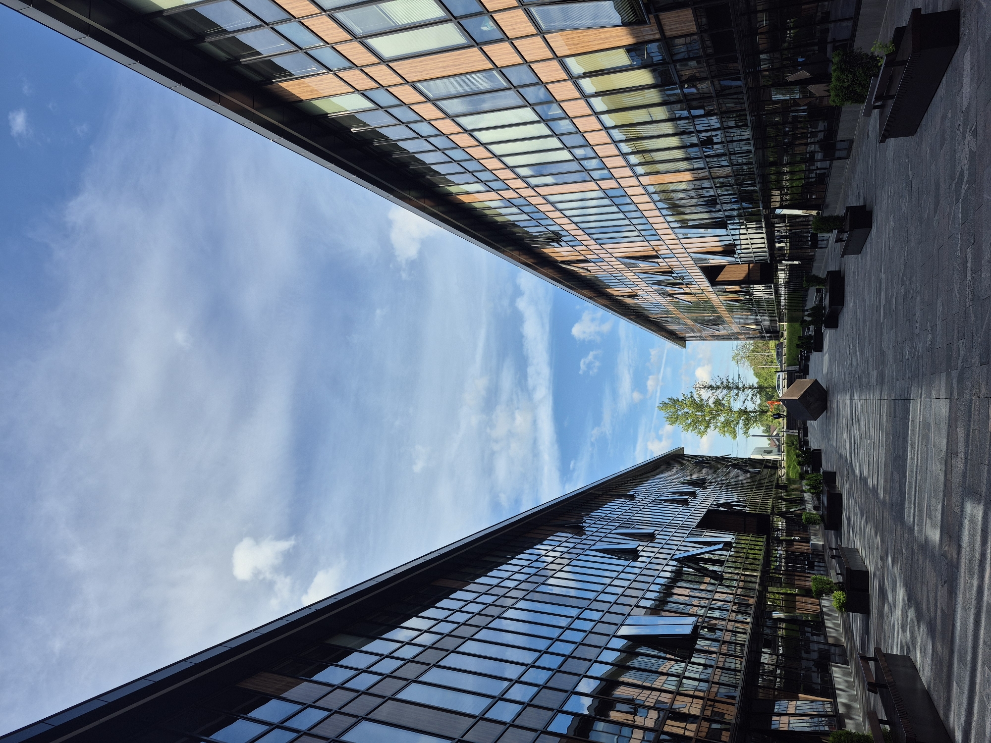 The image shows a low-angle, wide shot of modern architecture in Saint Petersburg, Russia. Two buildings, clad in glass and wood-toned panels, face each other, creating a narrow gap filled with blue sky and wispy white clouds. The sky appears bright, suggesting it is daytime, likely late morning or early afternoon, with the sun casting long shadows across the paved courtyard between the buildings. The architecture is sleek and contemporary, with clean lines and large windows that reflect the sky and surrounding structures.
The courtyard features dark, rectangular planters with some greenery, and a few dark benches. The paving stones are laid in a uniform pattern. In the distance, beyond the buildings, a few trees are visible, hinting at a park or green space nearby. There is no visible text in the image. The overall impression is one of urban modernity and a bright, clear day. The composition emphasizes the geometric shapes of the buildings and the contrast between the man-made structures and the natural elements of the sky and trees.